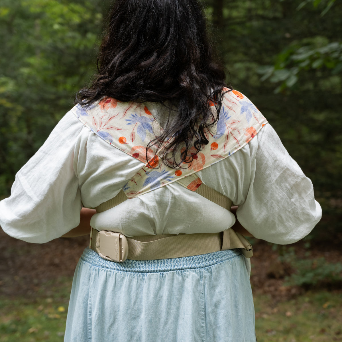 Back view of the Ambrosia Sprout Newborn Carrier, its printed panels crossed over the shoulders above a cream buckled waistband.