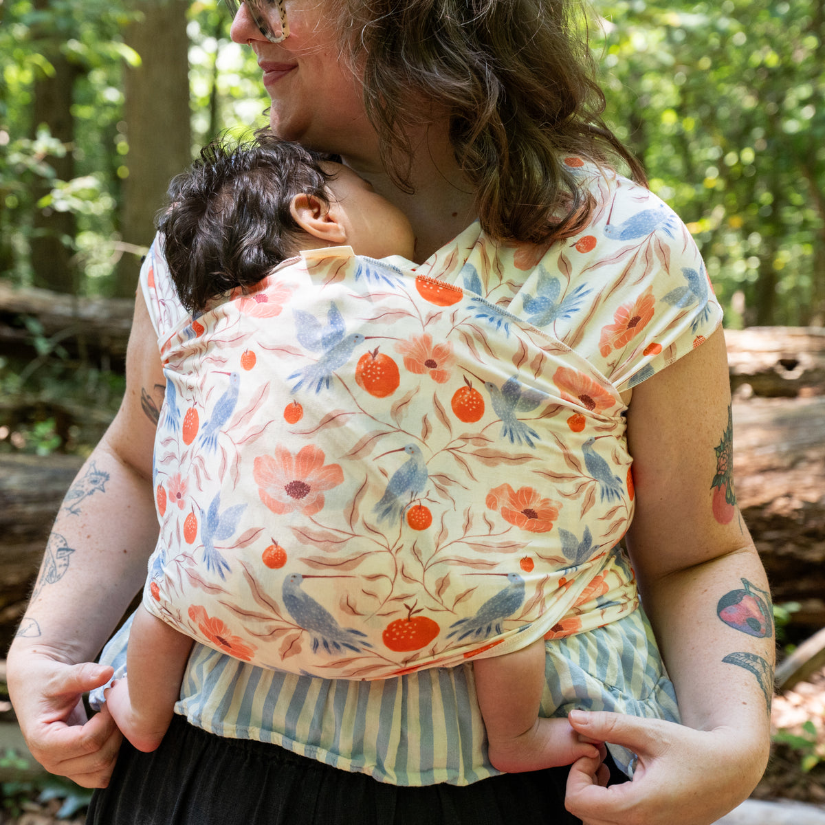 A wearer rests their head against a baby carried in the Ambrosia Baby Wrap, the printed stretchy fabric supporting the baby upright.
