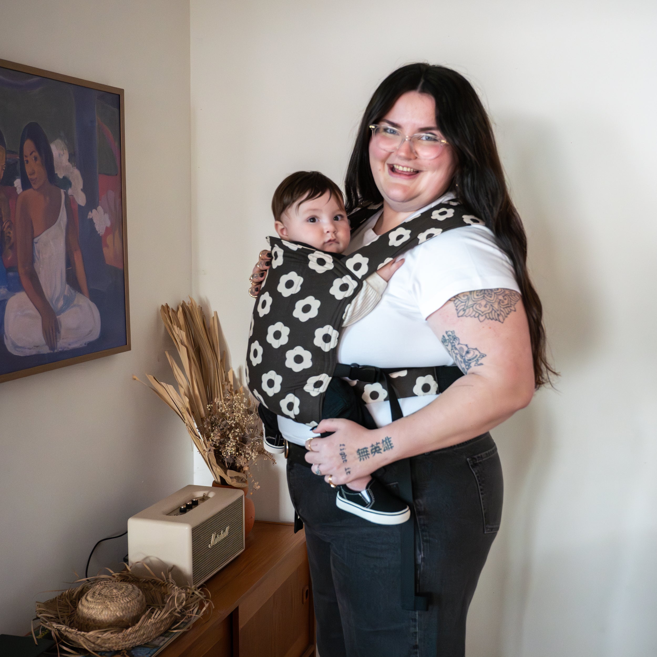 Woman holding a baby in a black and white floral baby buckle carrier indoors.