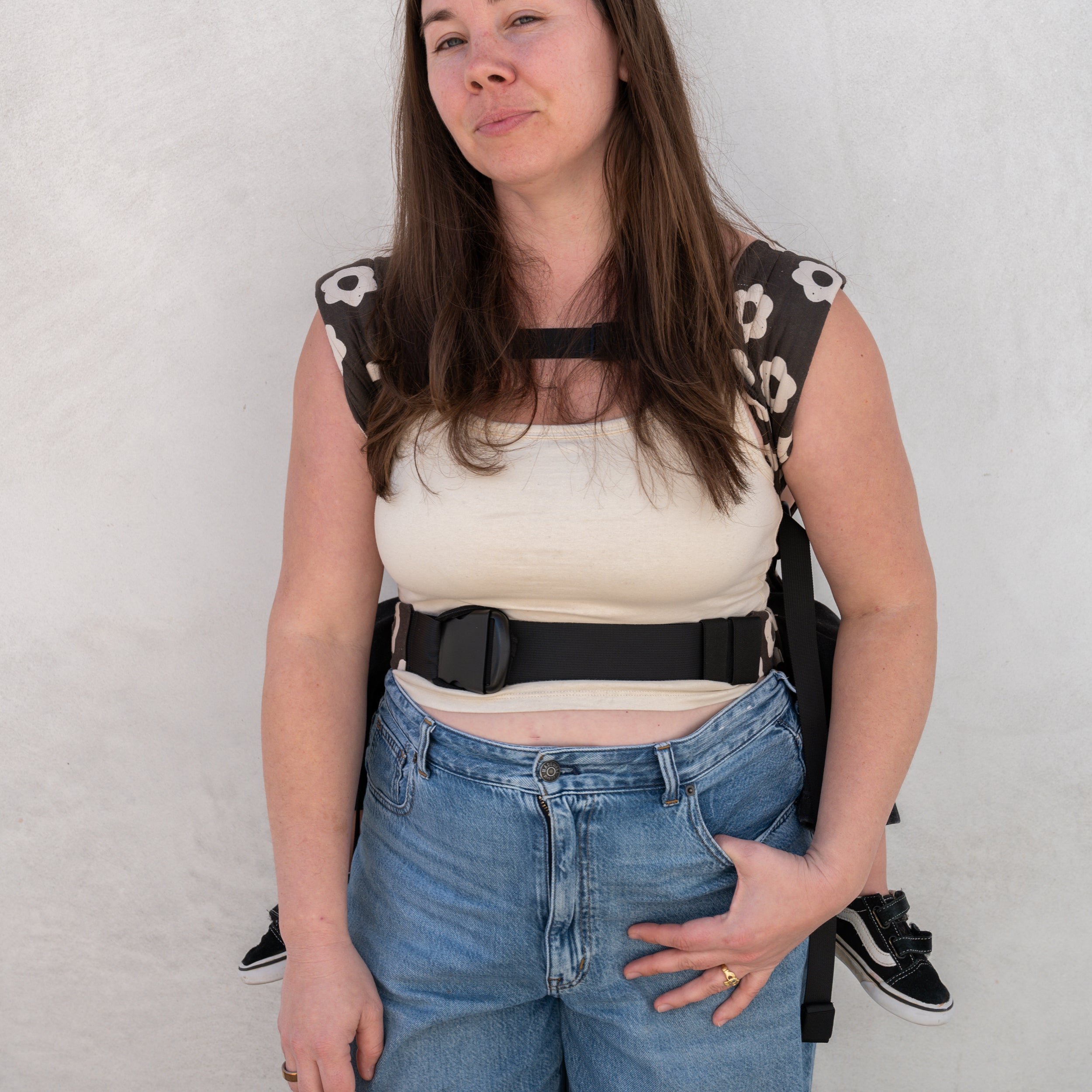 Woman wearing a black and white floral buckle carrier over a beige top and blue jeans against a white background