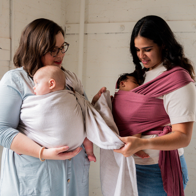 White mother wearing gray ring sling baby carrier and Latina mother wearing newborn in pink baby wrap carrier. 