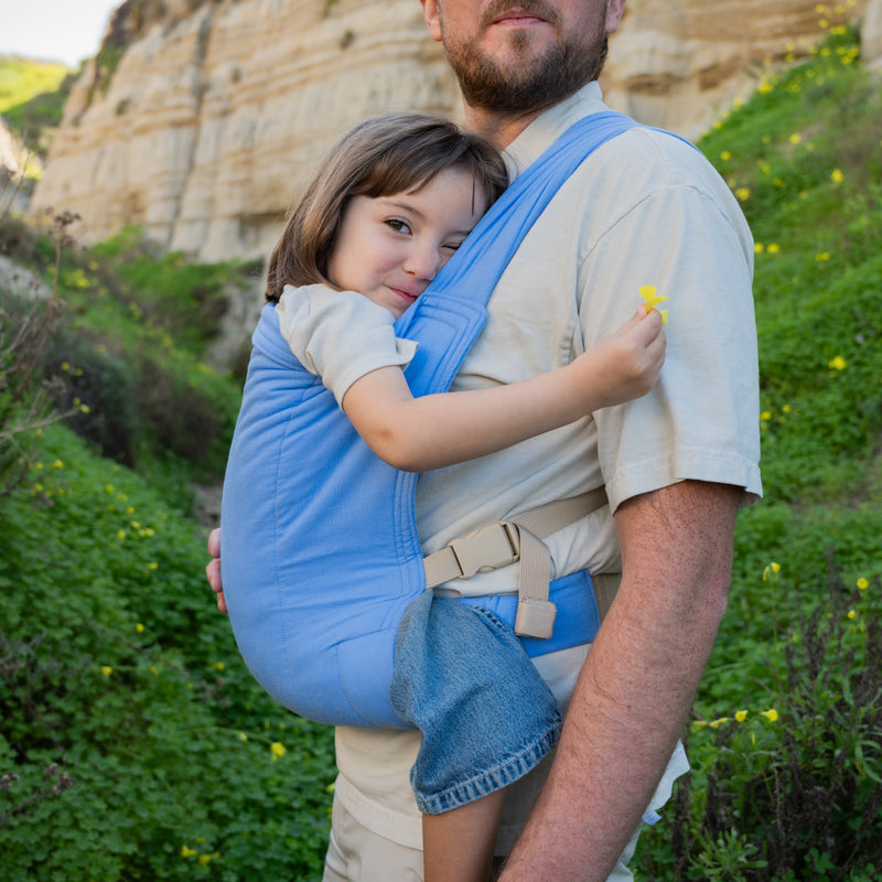 Man carrying a child in a blue baby carrier outdoors with greenery and rocks in the background