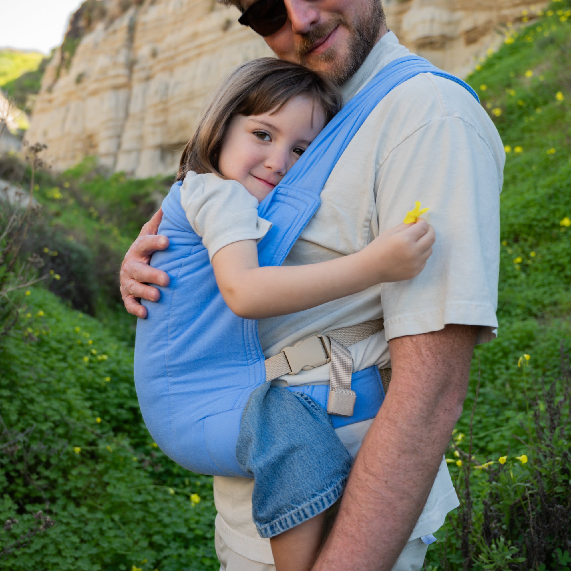 Man carrying a child in a blue baby carrier outdoors with greenery and rocks in the background