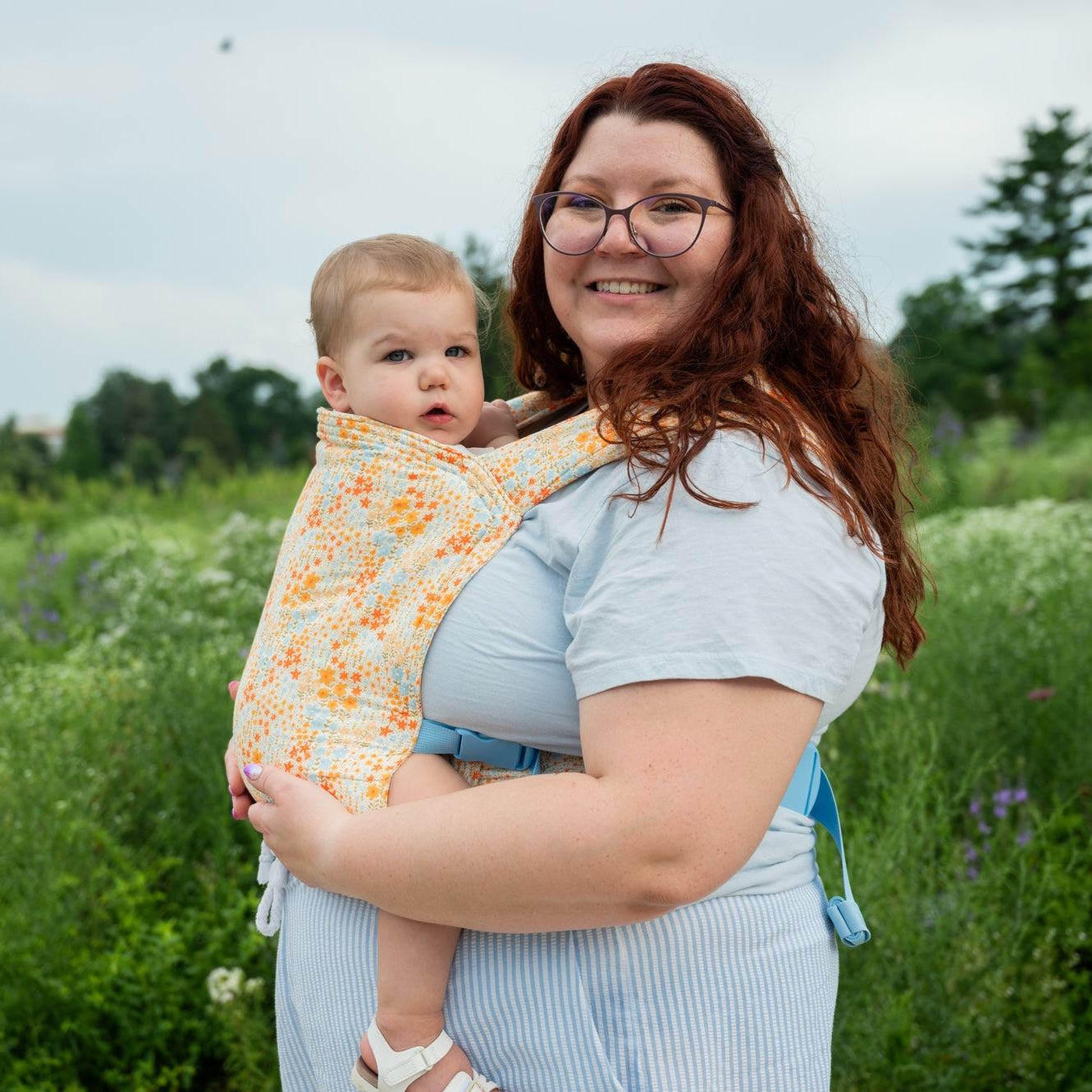 Woman holding a baby in a carrier outdoors in a field with greenery and blue sky.