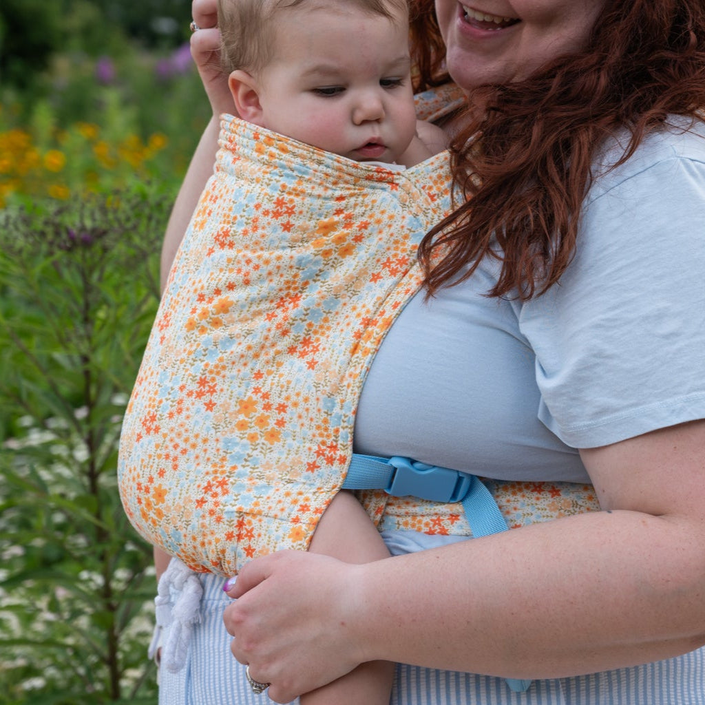 Woman holding a baby in a floral sling amidst a natural setting with flowers and greenery.
