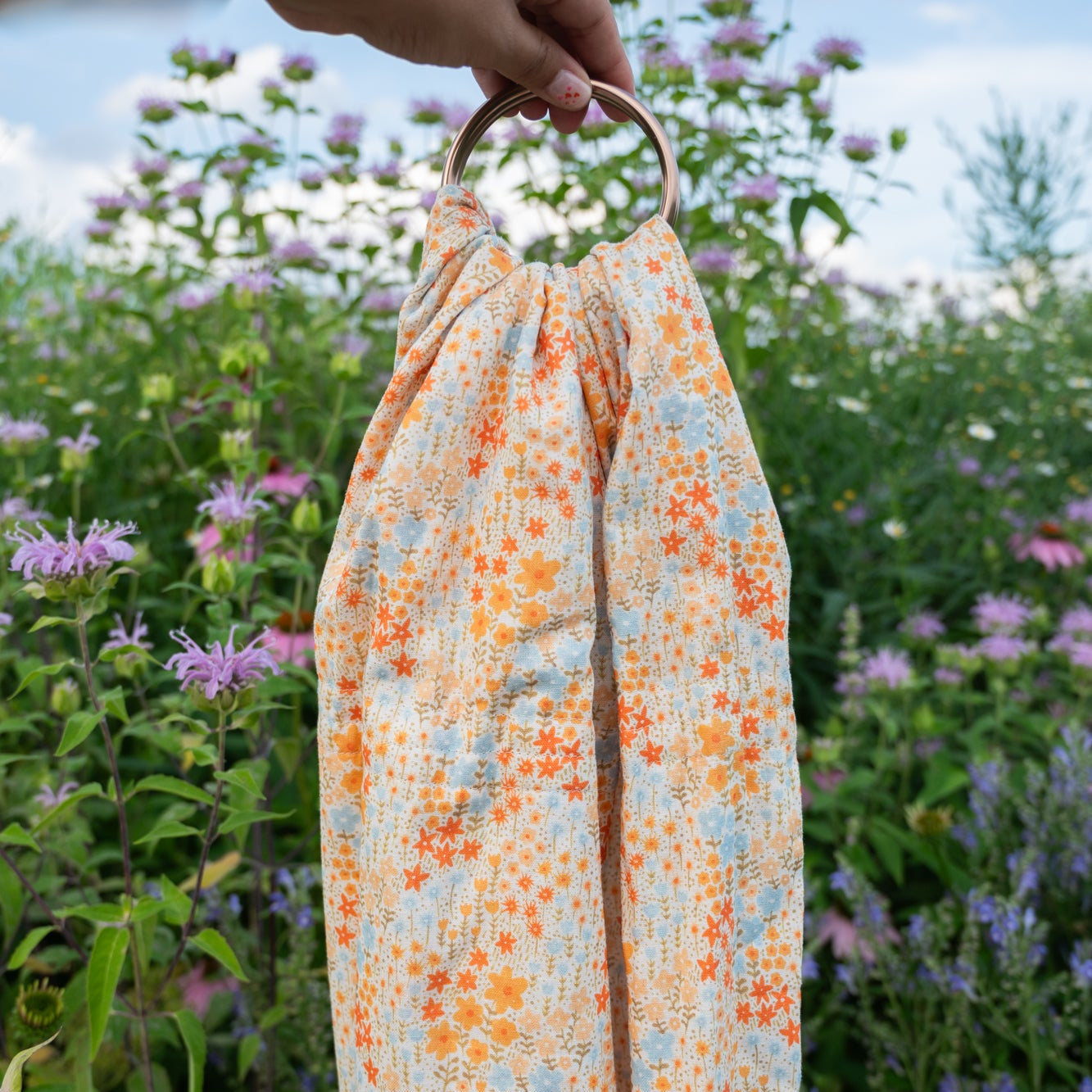 Floral-patterned ring sling carrier held by a hand with a natural background of flowers and greenery