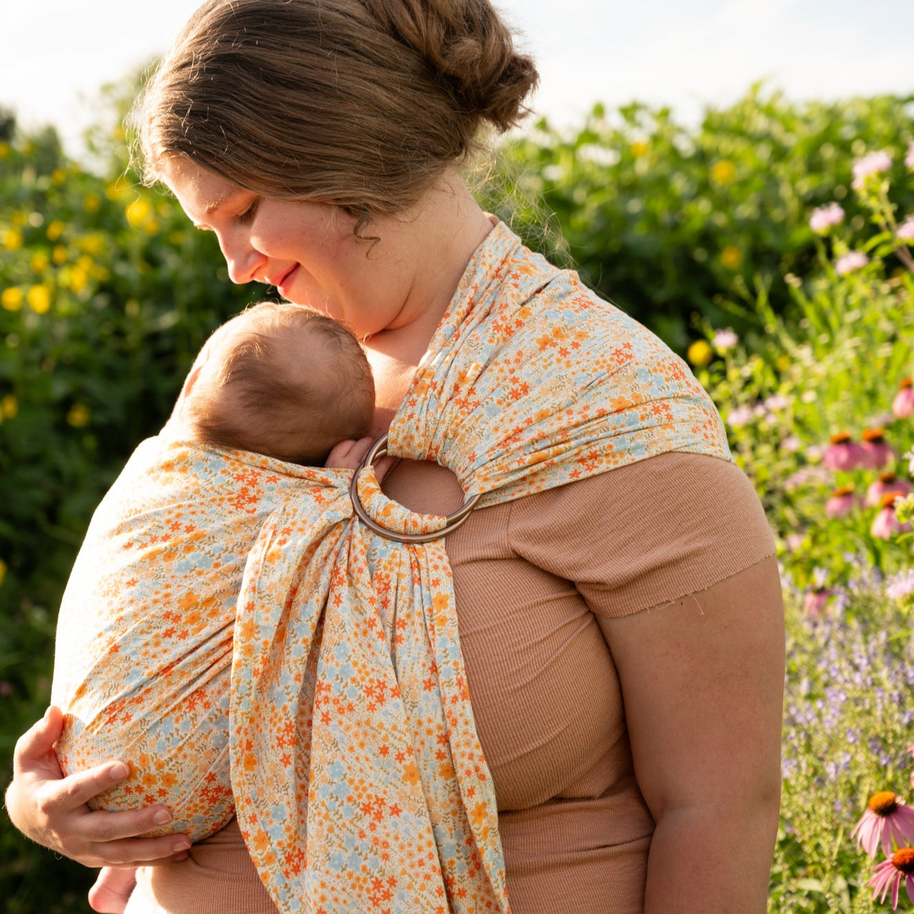 Woman wearing newborn in floral print ring sling with a greenery background. 