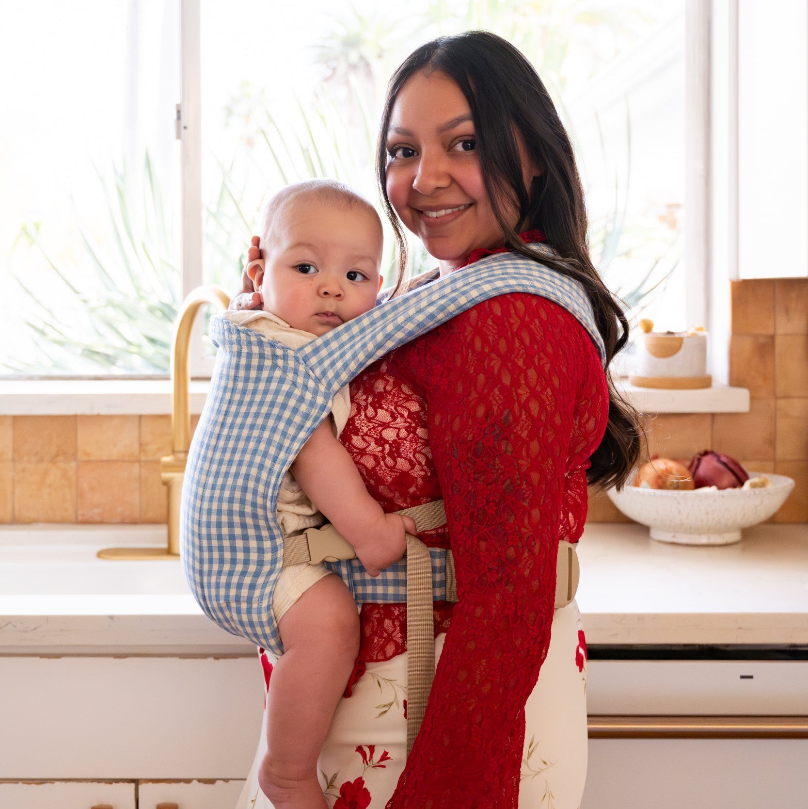 Woman holding a baby in a blue gingham buckle baby carrier in a kitchen