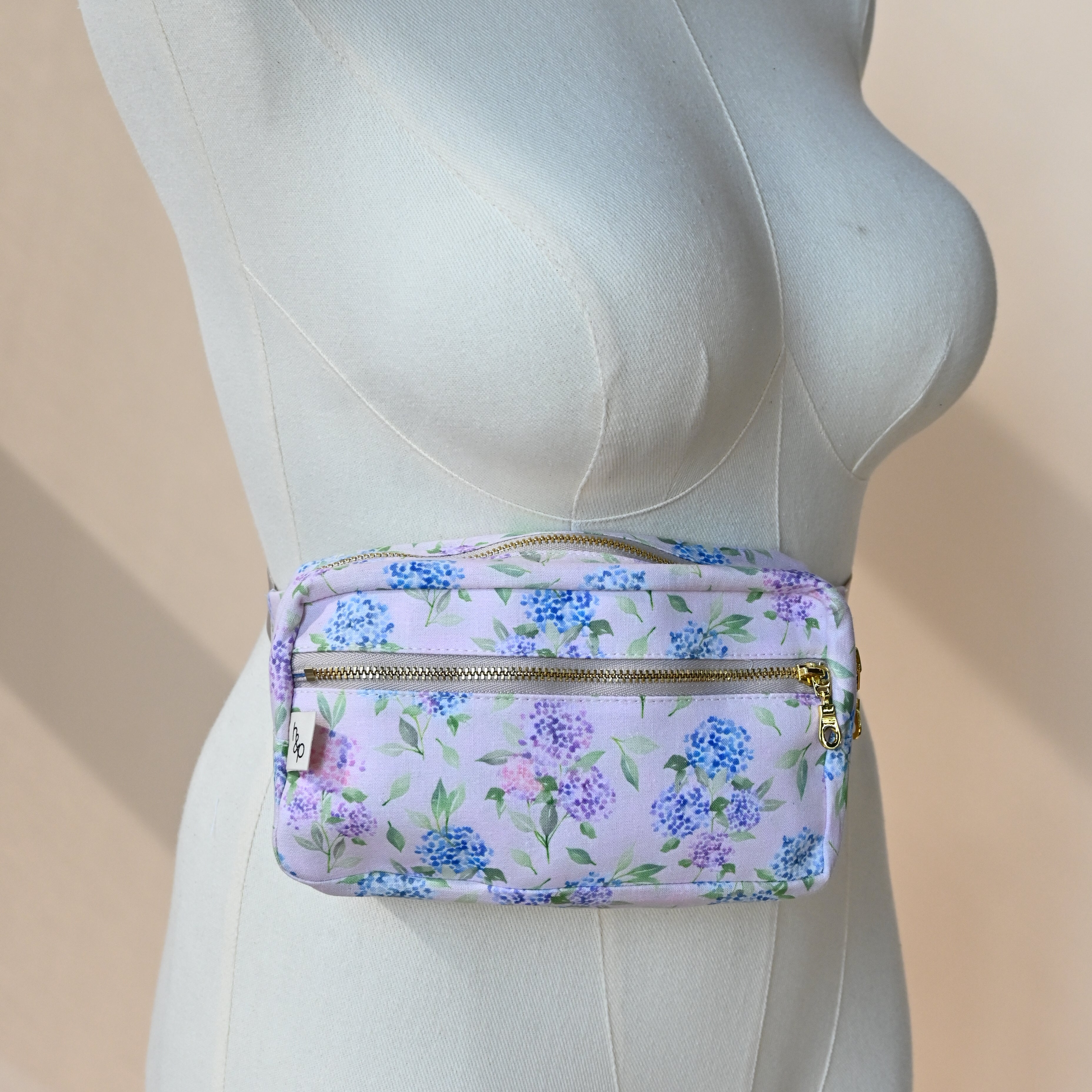 Fanny Pack in Blush — hydrangea-print belt bag with gold zips shown on a dress form, blue and lavender blooms on a soft pink background.