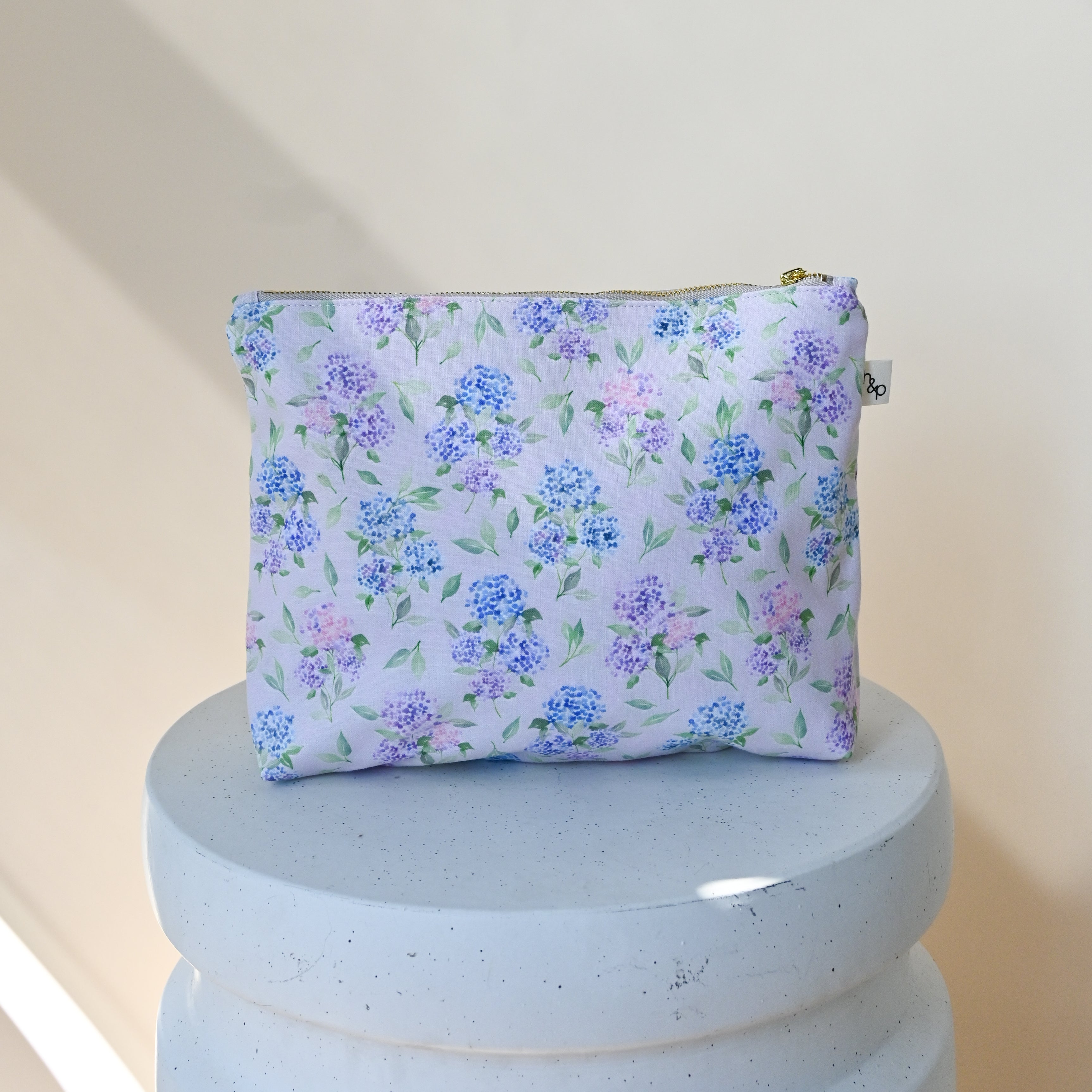 Large Flat Bottom Bag in Blush — the zippered pouch shown on a pedestal, hydrangea clusters in blue, lavender and pink on a soft pink background.