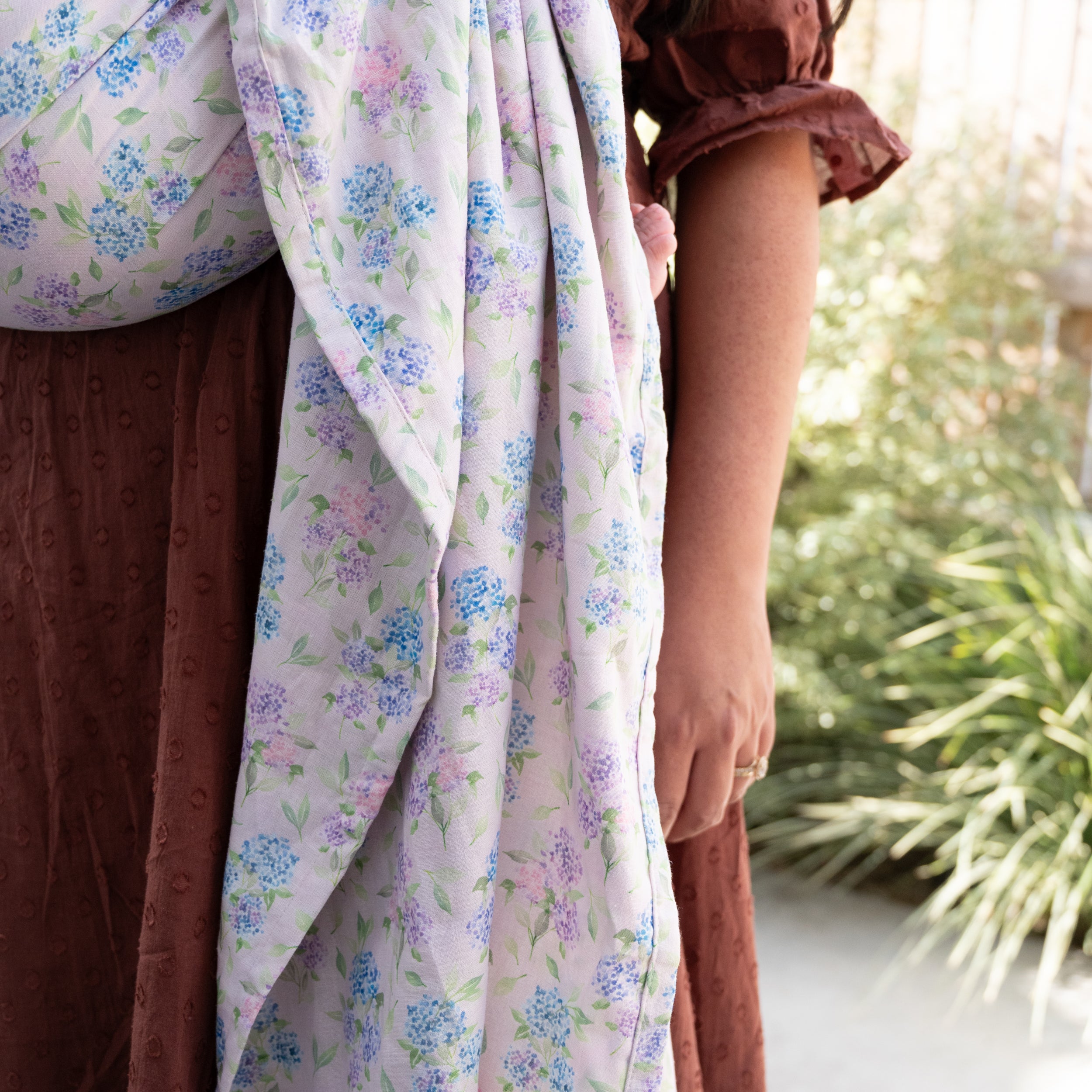 Ring Sling in Blush — the printed tail draped down the wearer's side, hydrangea blooms in blue, lavender and pink on a soft pink background.