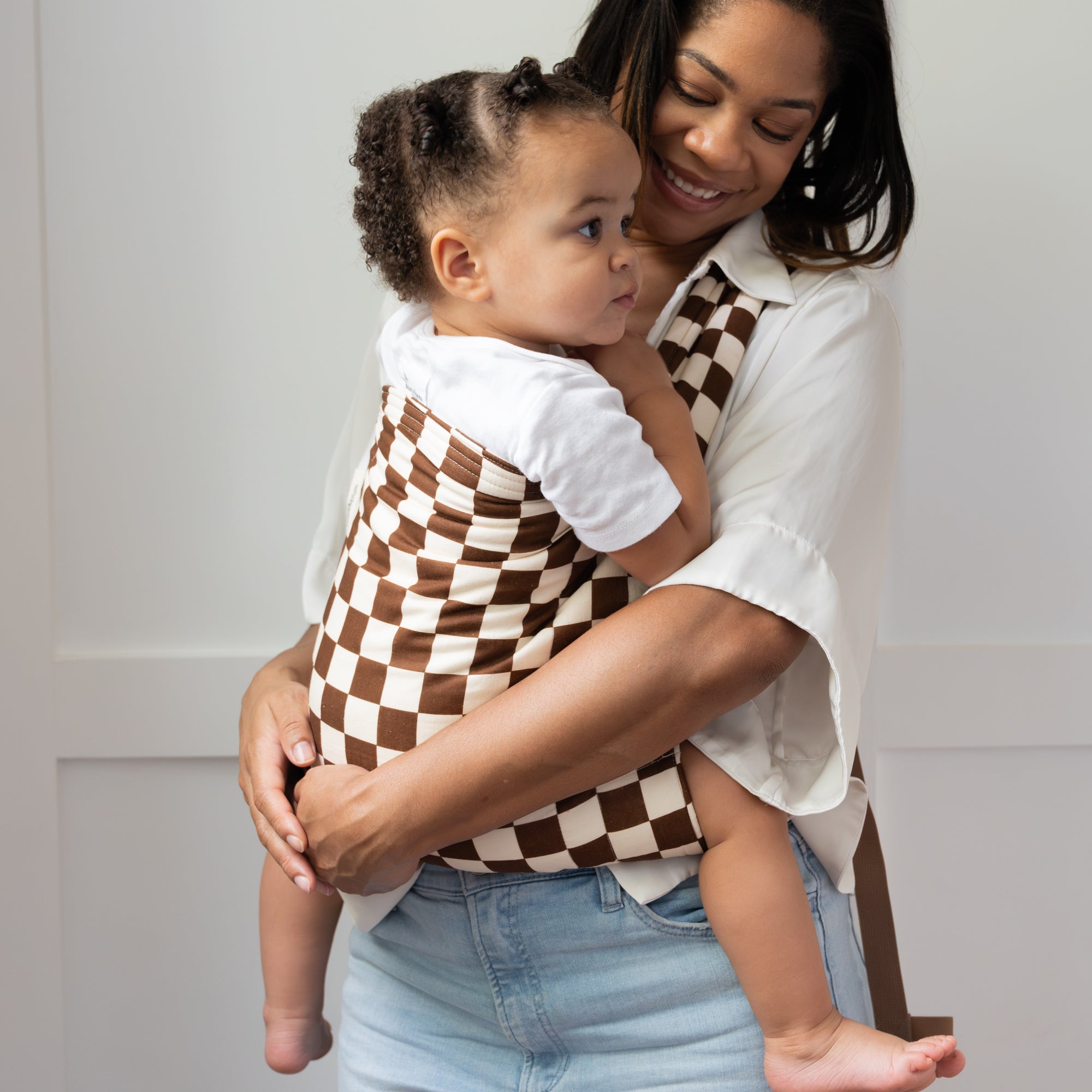 Woman holding a child in a brown and white checkered baby carrier against a neutral background