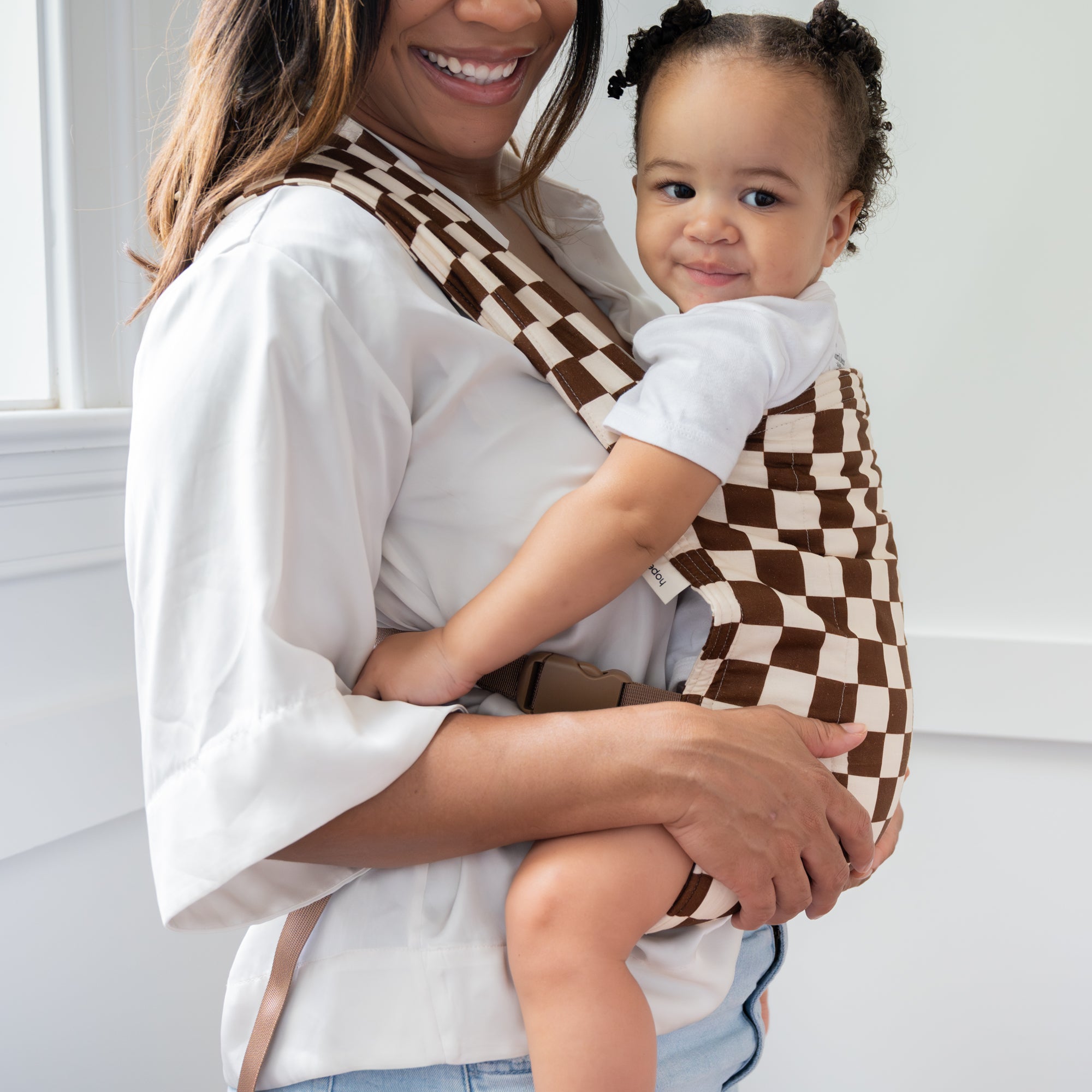 Woman holding a child in a brown and white checkered baby carrier against a neutral background