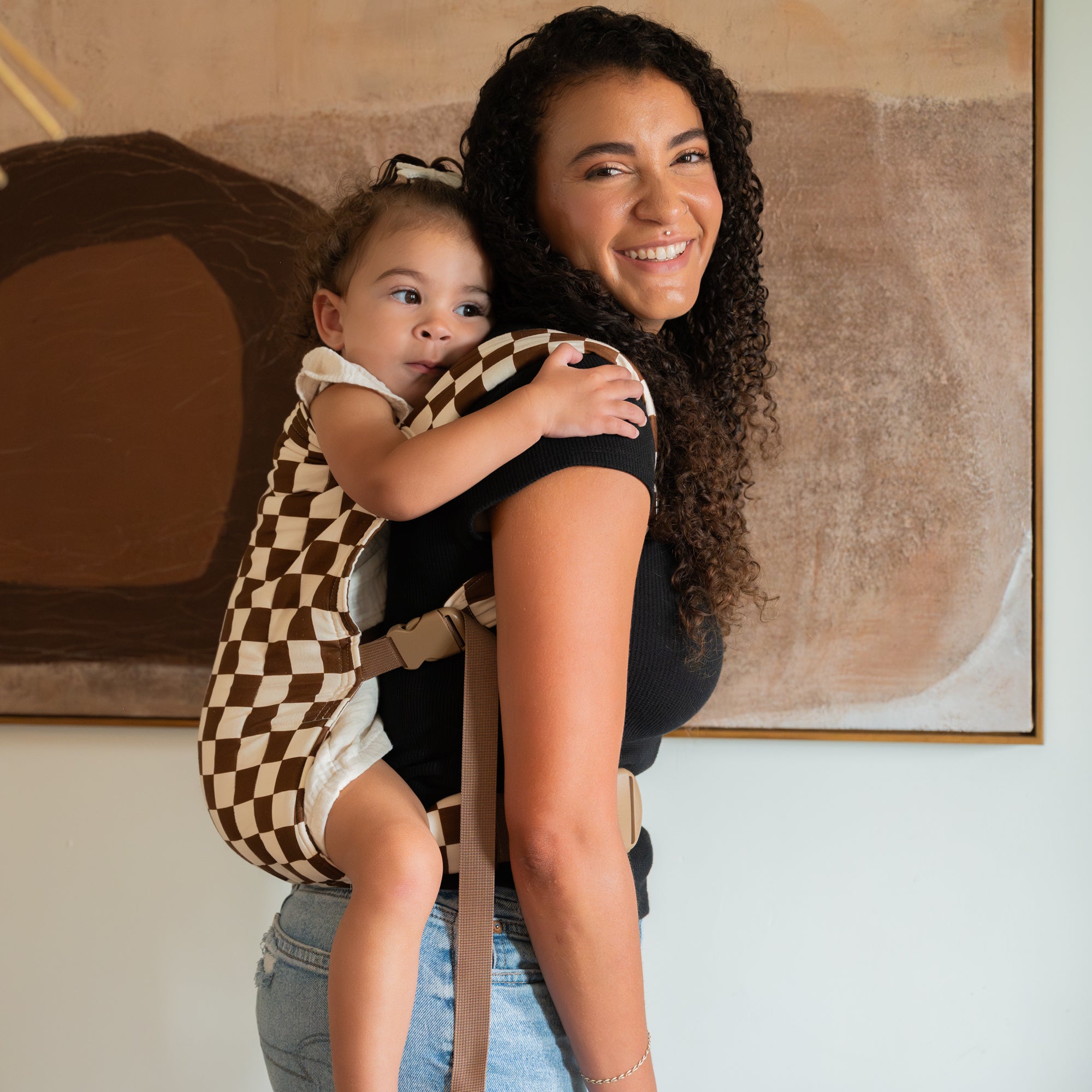 Woman holding a child in a brown and white checkered baby carrier against a neutral background