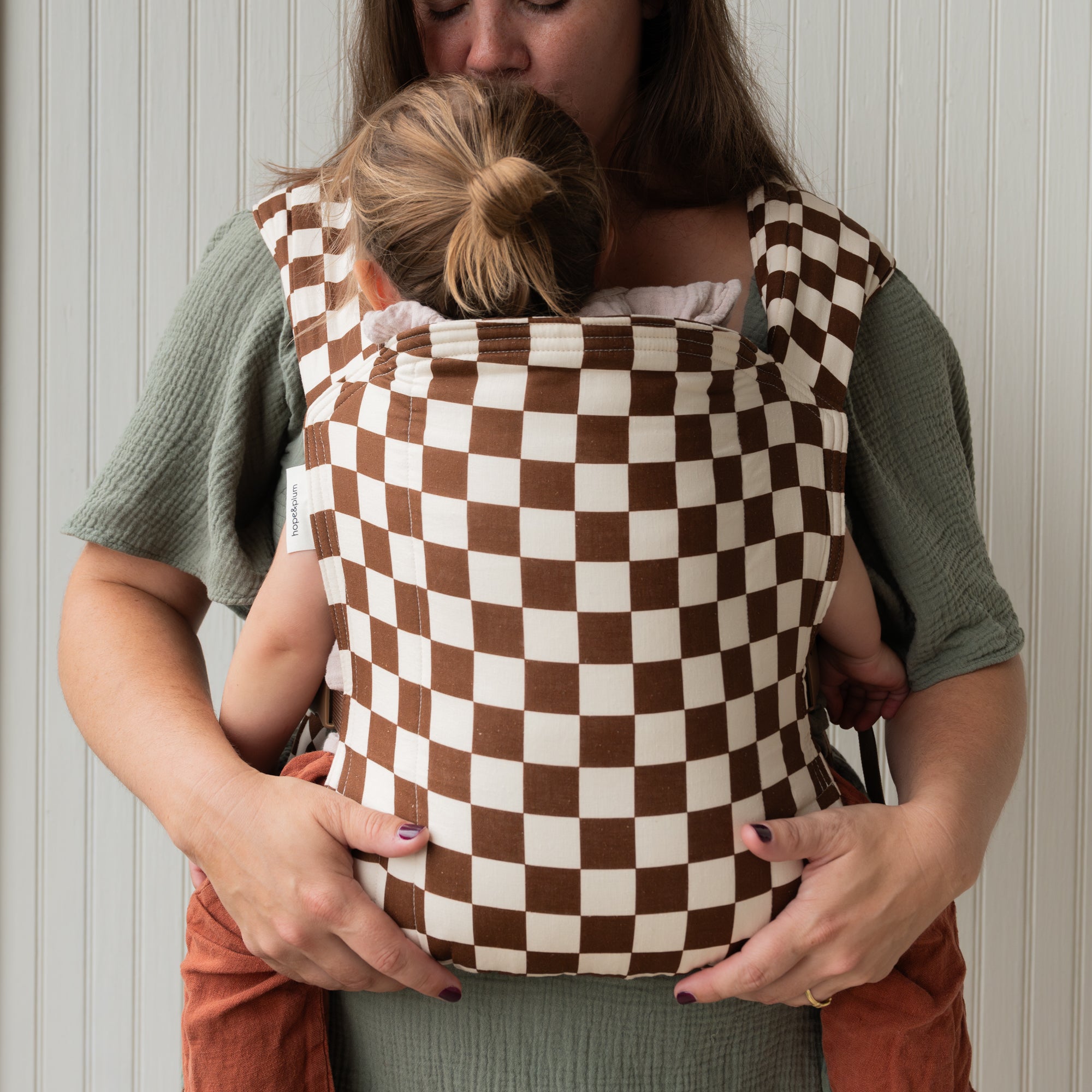 Woman holding a child in a brown and white checkered baby carrier against a neutral background