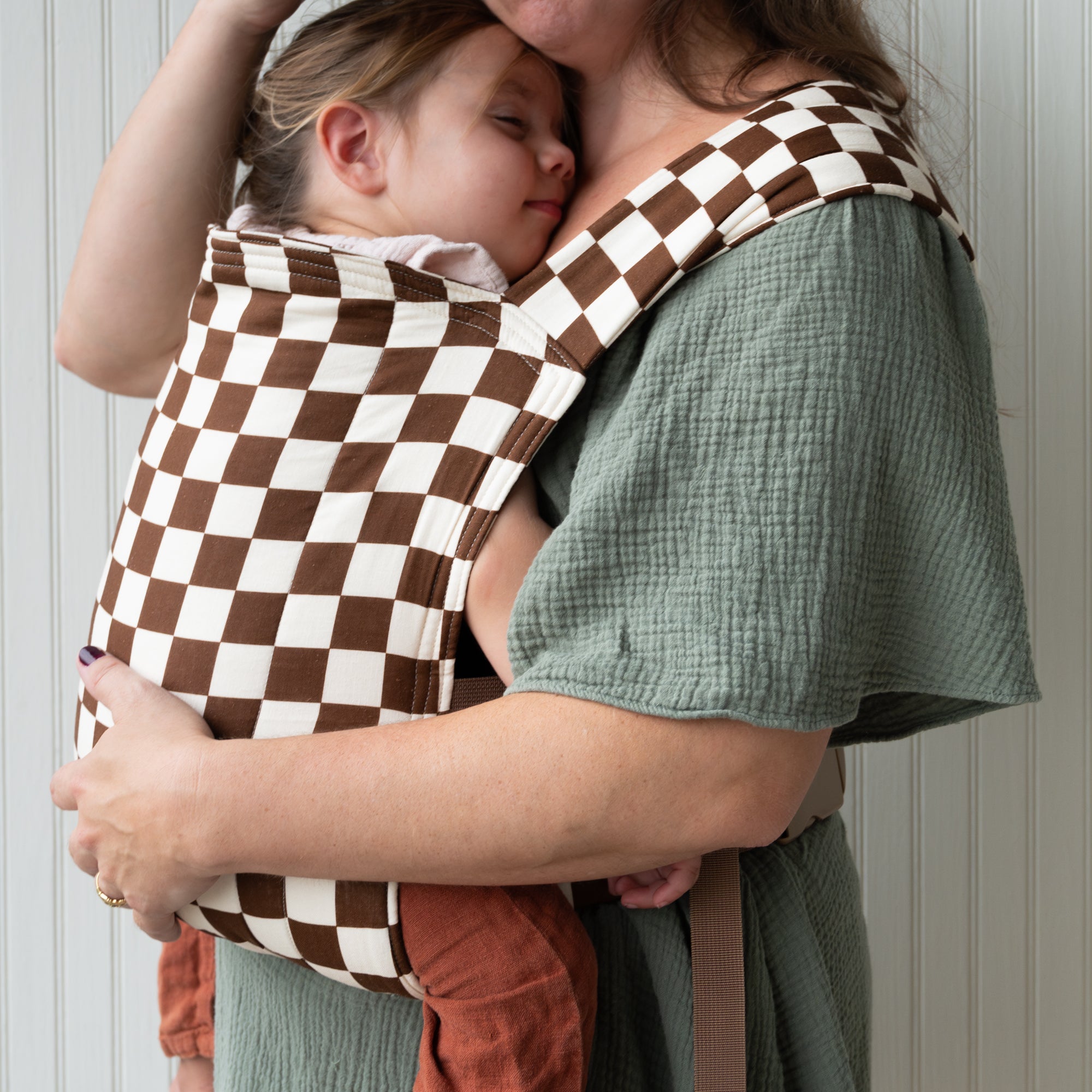 Woman holding a child in a brown and white checkered baby carrier against a neutral background