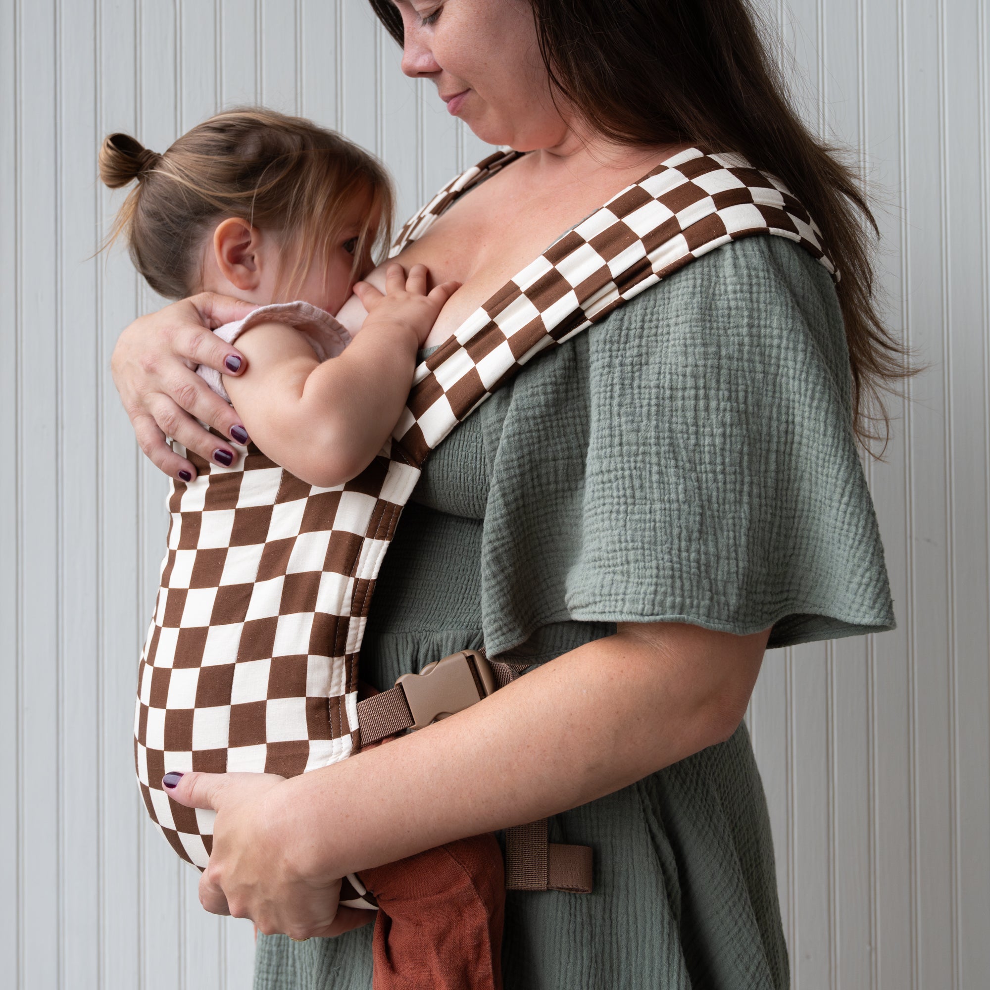 Woman holding a child in a brown and white checkered baby carrier against a neutral background