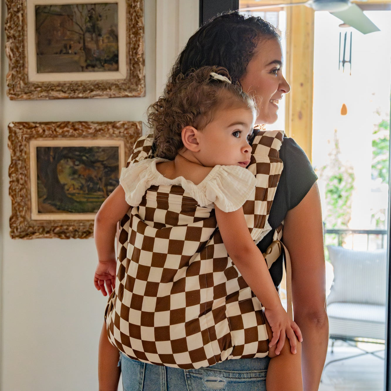 Woman carrying a child in a checkered toddler carrier inside a home.