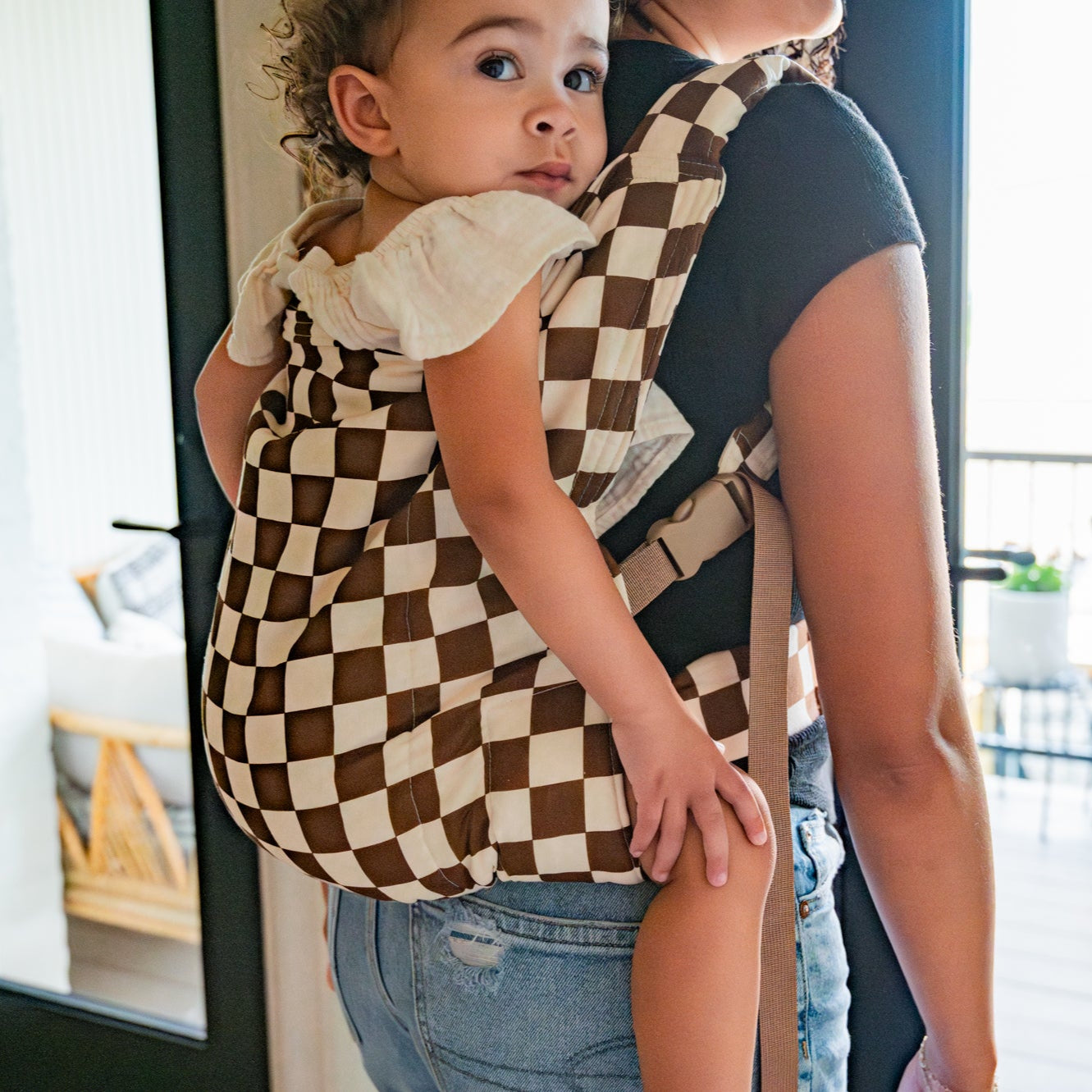 Woman carrying a child in a checkered baby carrier indoors.