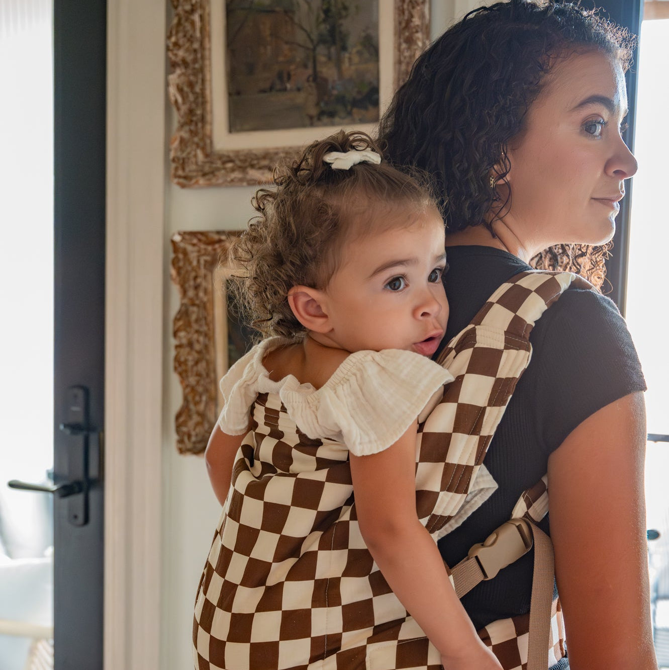 Woman carrying a child in a checkered kid buckle carrier inside a home.