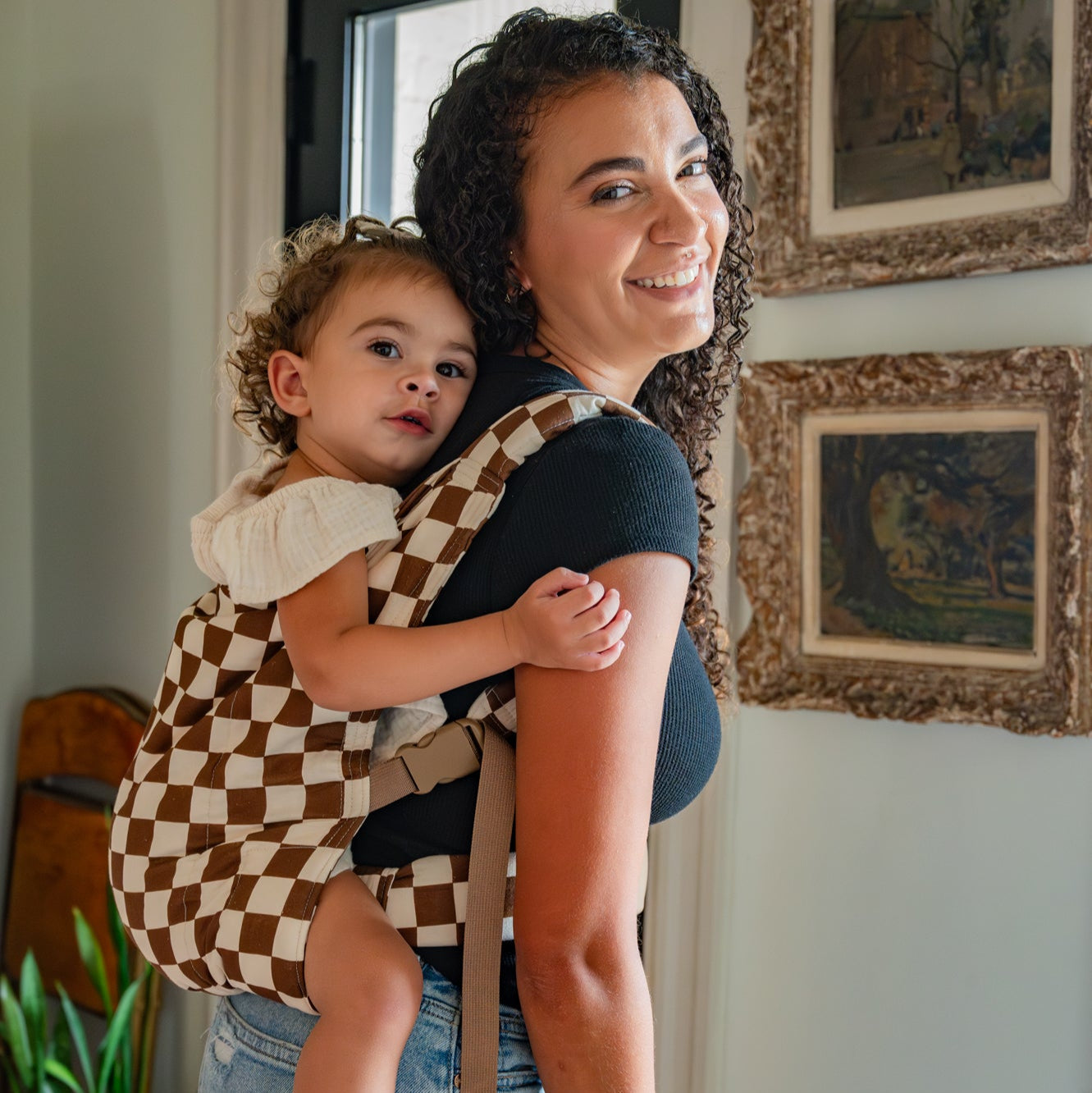 Woman holding a child in a checkered baby carrier indoors.
