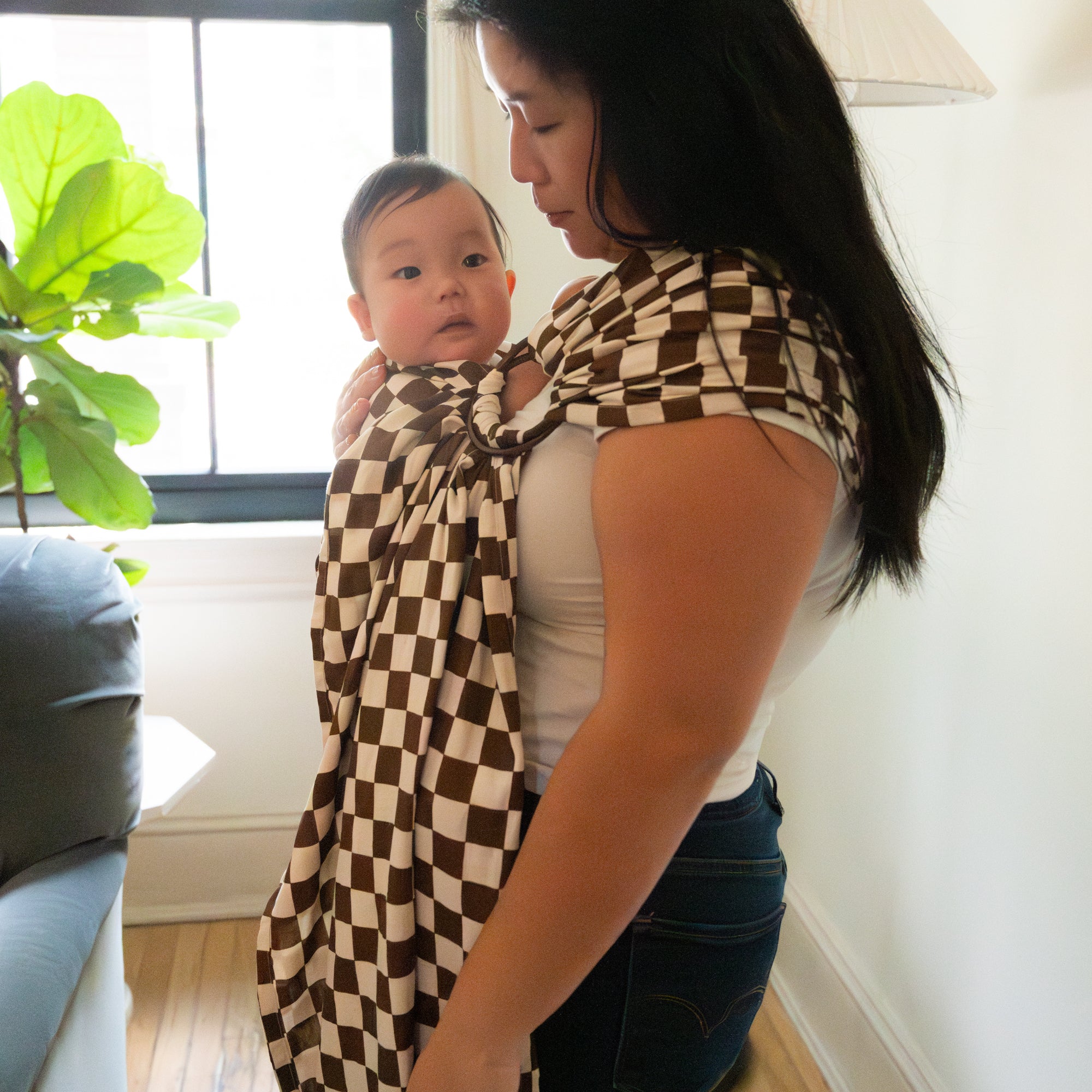 Woman holding a baby wrapped in a checkered ring sling carrier in a home setting