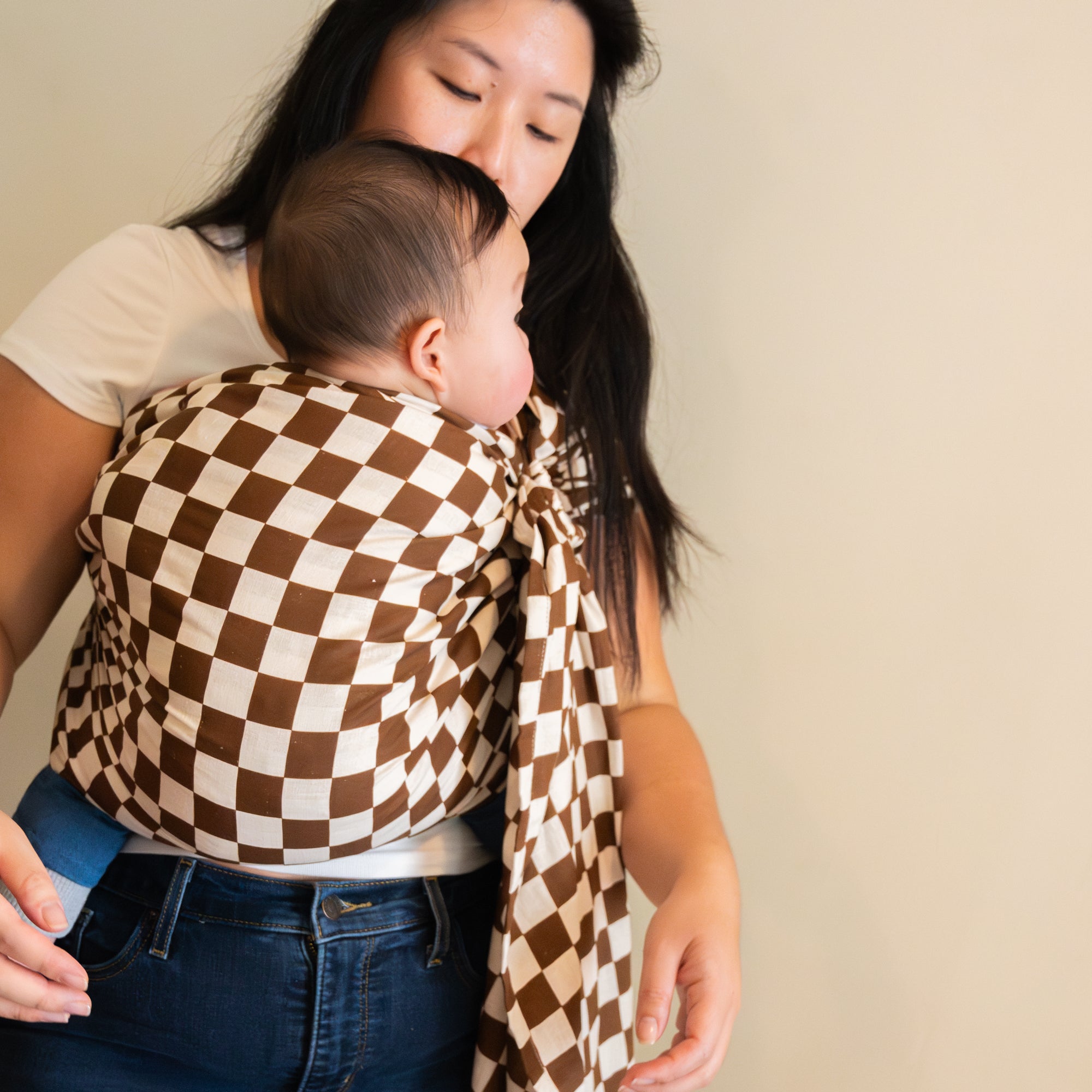 Woman holding a baby wrapped in a brown and white checkered ring sling carrier against a beige background