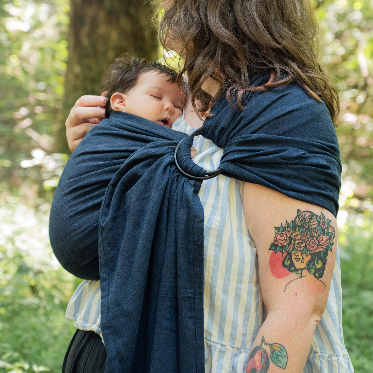 A wearer leans down to a sleeping baby carried in the Caspian Ring Sling, the dark indigo chambray drawn across the body to the rings.