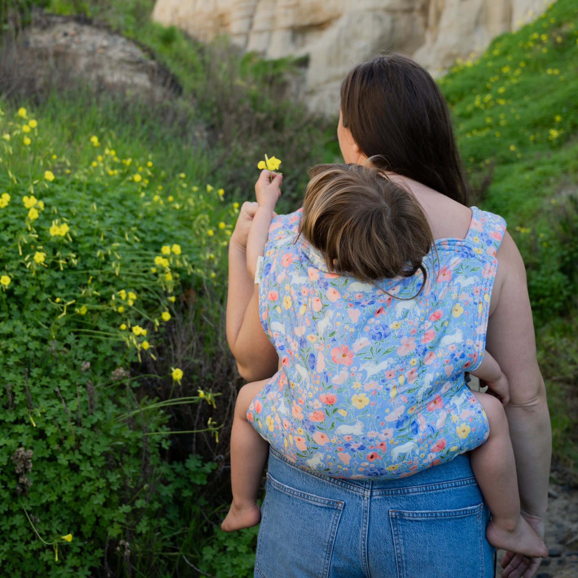 Woman holding a child in a floral unicorn baby carrier amidst greenery and rocks.