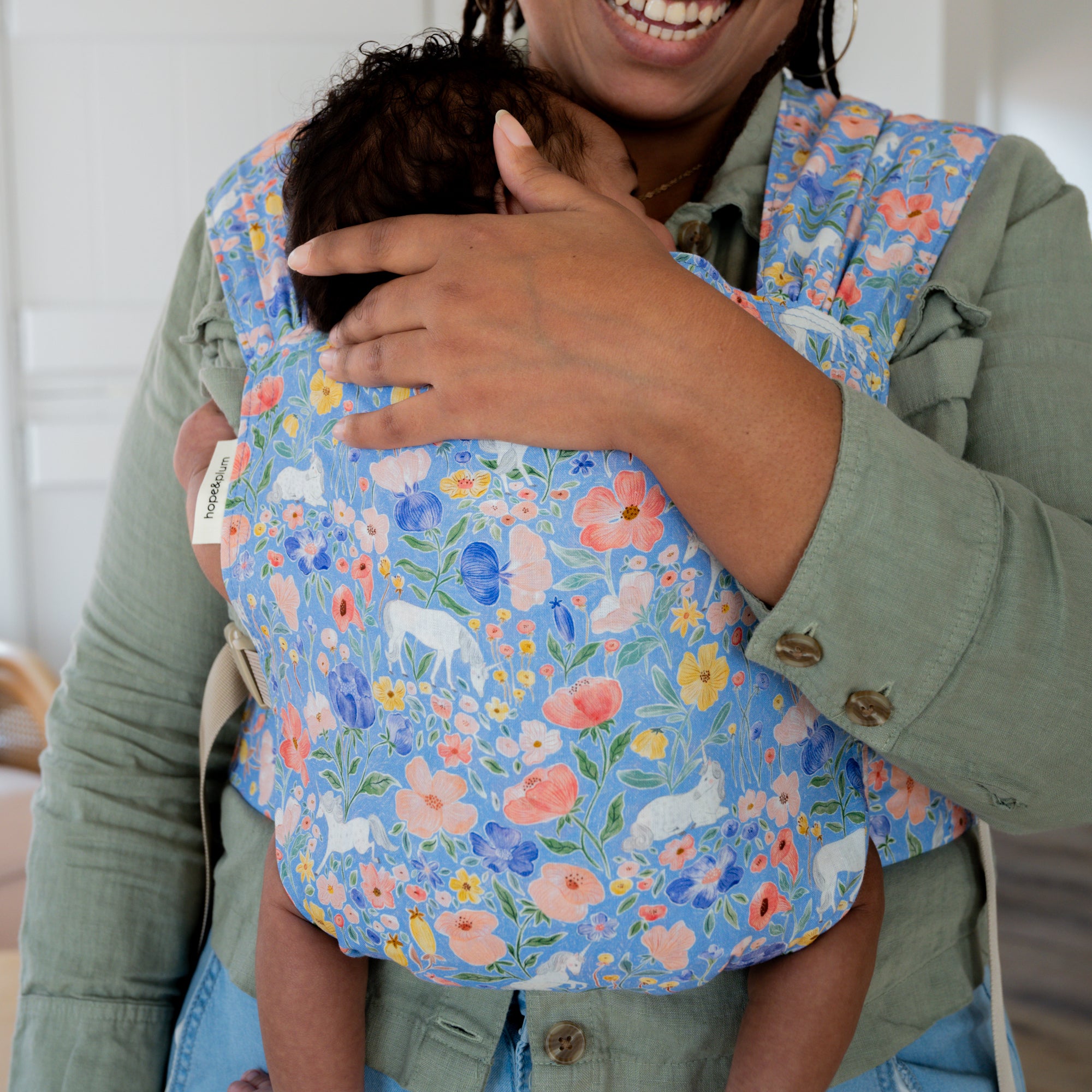 Person wearing a floral unicorn baby carrier with a child inside, smiling.