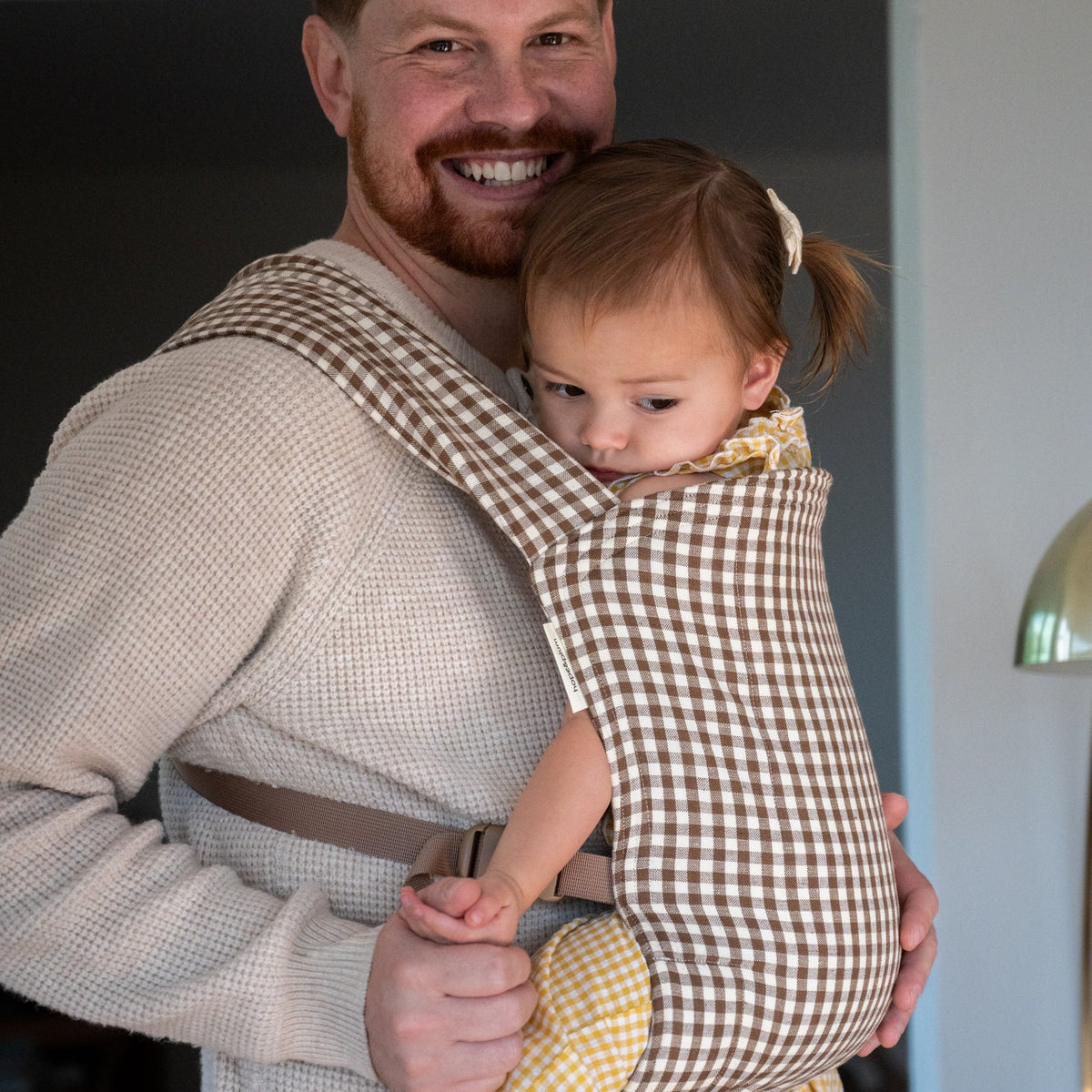 Lark Baby Carrier in Clove — caregiver smiling with a toddler nuzzled into his chest, brown-and-cream gingham carrier, at home.