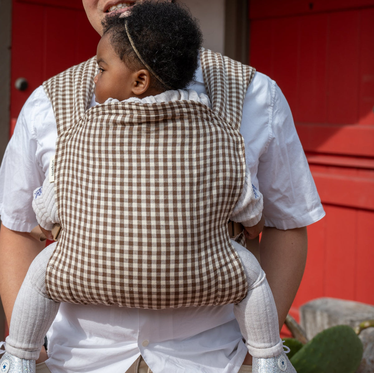 Lark Baby Carrier in Clove — back view of the full gingham body panel, caregiver standing on a porch with a red door and potted cactus.