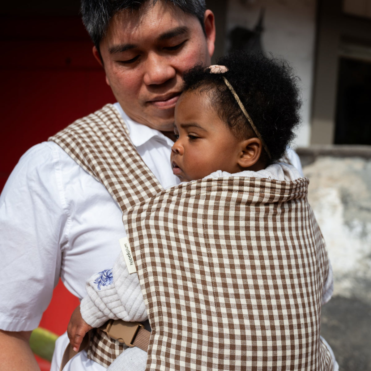 Lark Baby Carrier in Clove — caregiver smiling down at a toddler tucked into the gingham carrier, cactus and a red door behind them.
