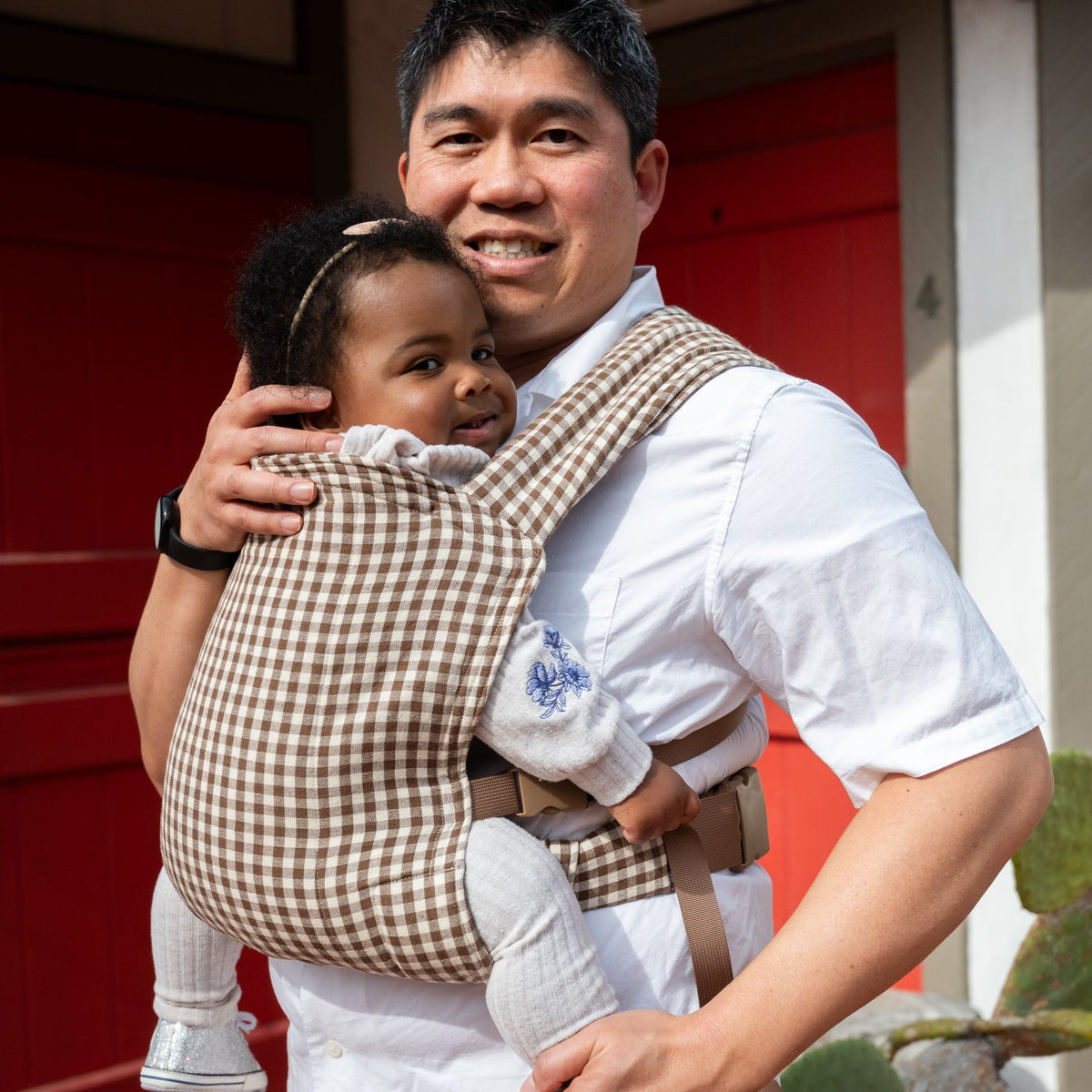 Lark Baby Carrier in Clove — caregiver embracing a toddler front-carried in the gingham carrier, red door and cactus in the background.