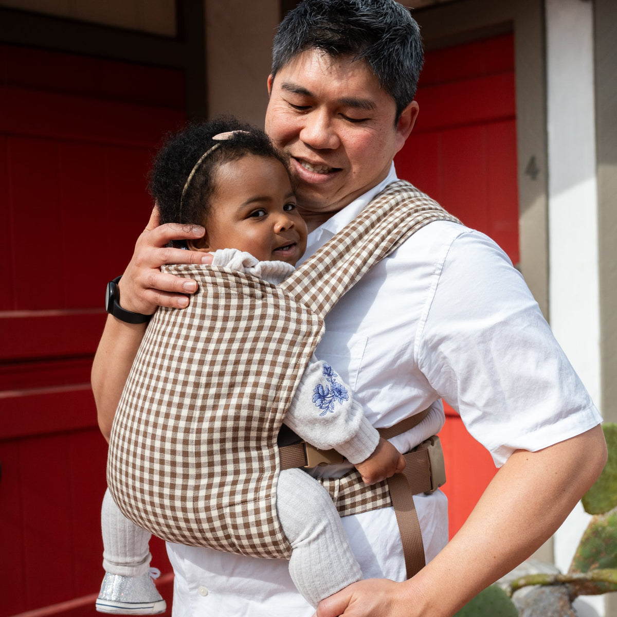 Lark Baby Carrier in Clove — caregiver smiling down at a toddler in the gingham carrier, sparkly shoes dangling below the seat.