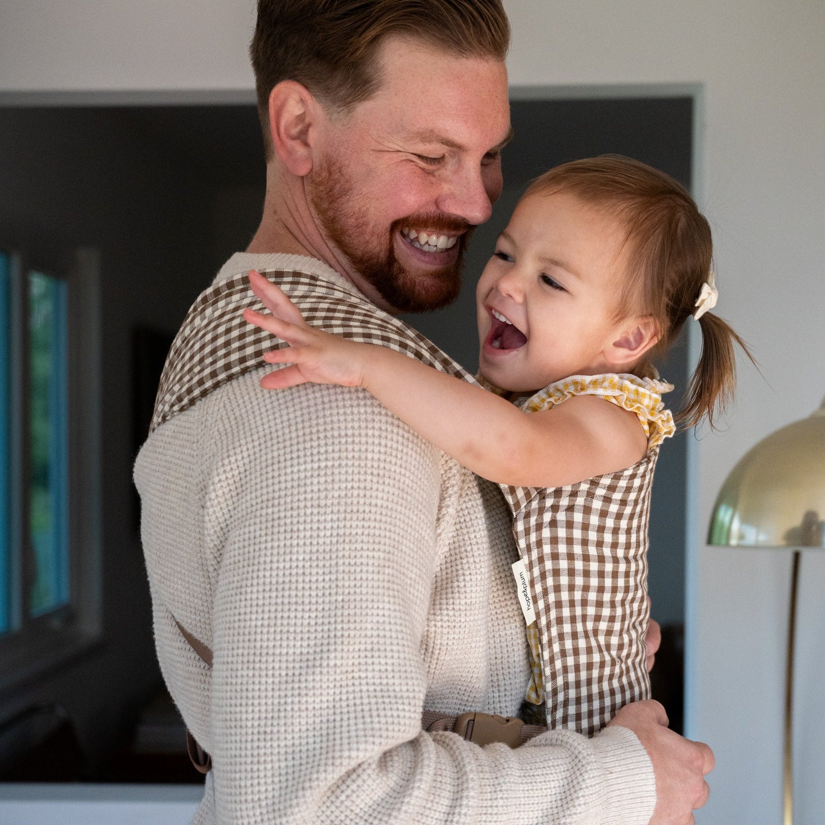 Lark Baby Carrier in Clove — caregiver laughing with a toddler reaching out an arm, gingham carrier visible at his shoulder, at home.