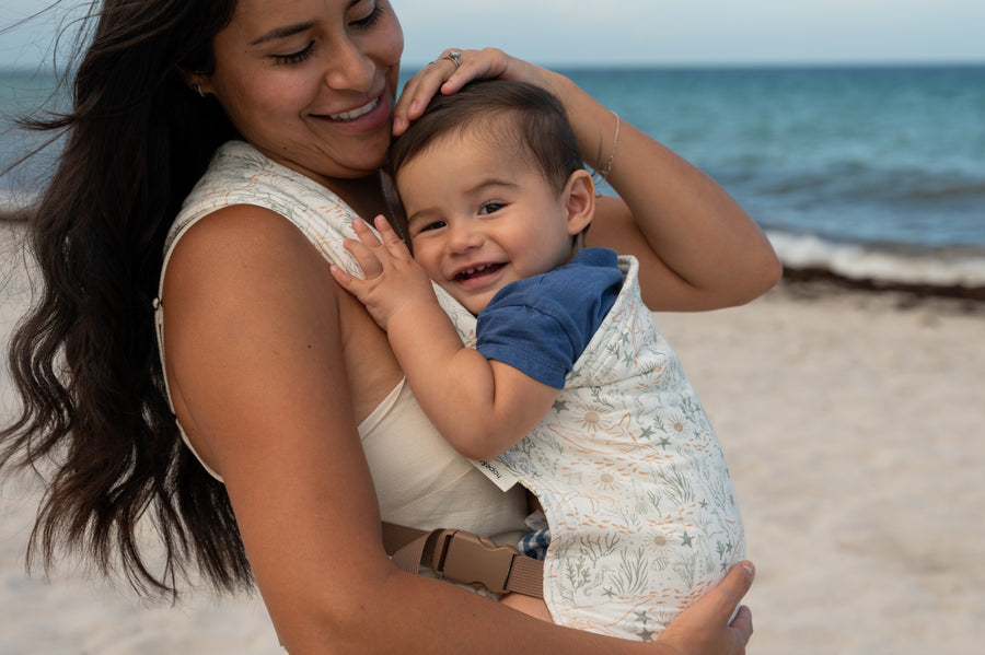 A smiling caregiver holds a grinning baby in the Cove Lark — a cream woven fabric with delicate sea creatures and coral in soft sage and terracotta — on a breezy sandy beach.