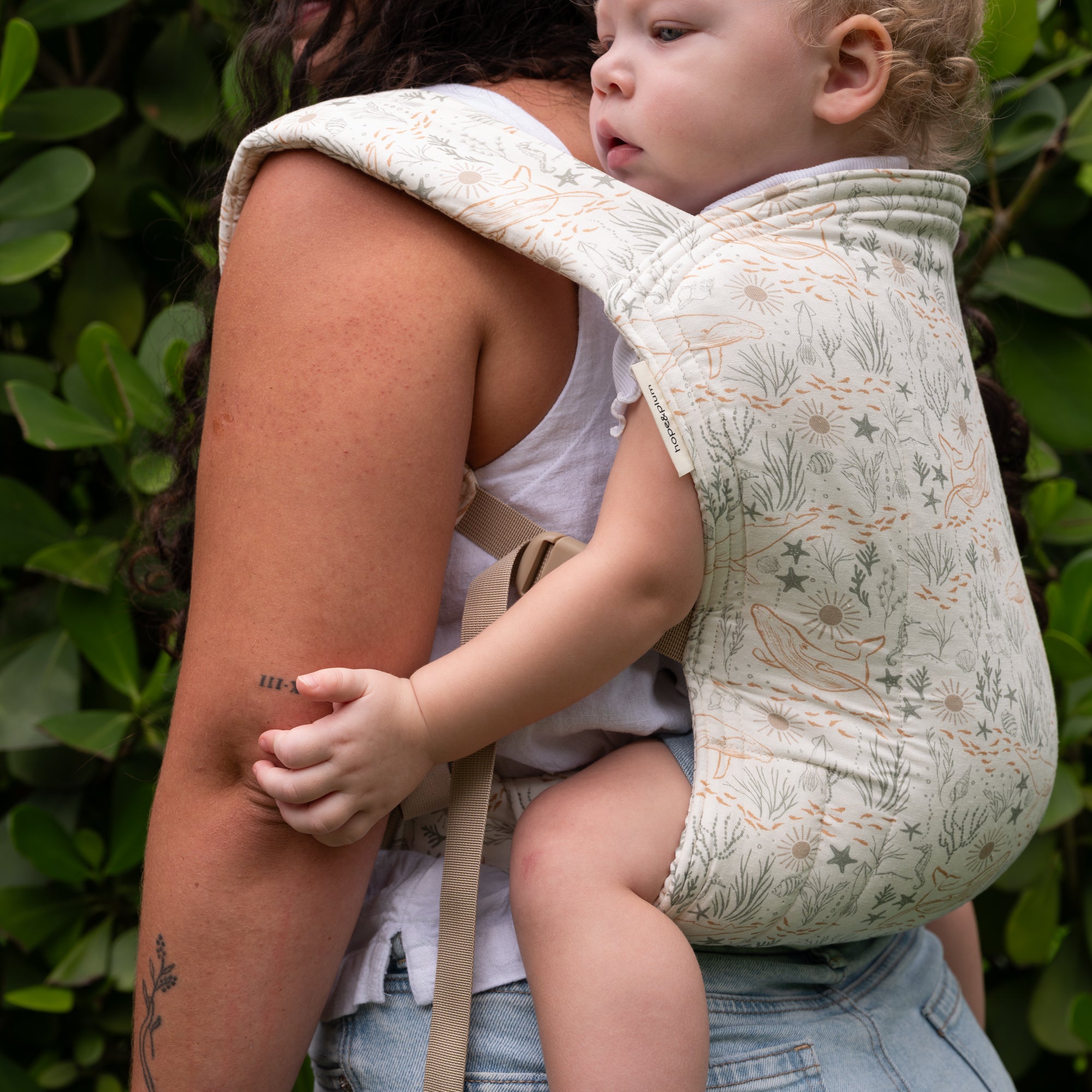 White Lark Baby Carrier with an illustrated sea-life print, in the Cove colorway, worn back-carry in a garden