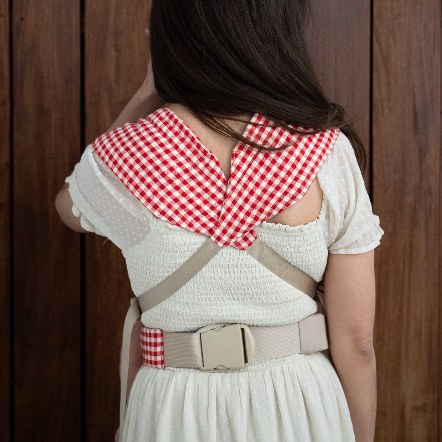 The back of a person wearing a white dress with a red and white gingham carrier straps over the shoulders against a wooden background
