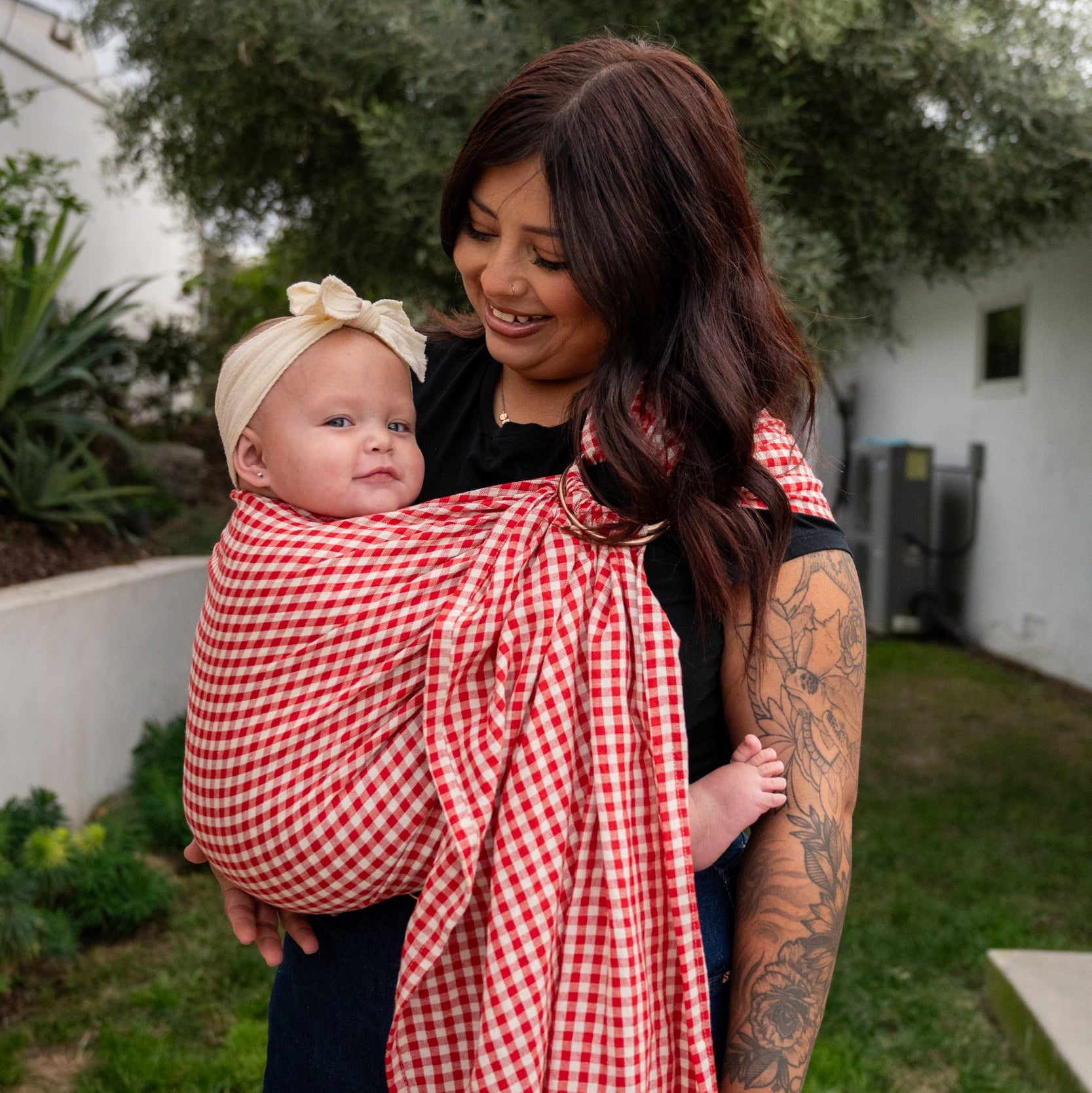 Woman holding a baby wrapped in a red and white gingham ringsling outdoors.