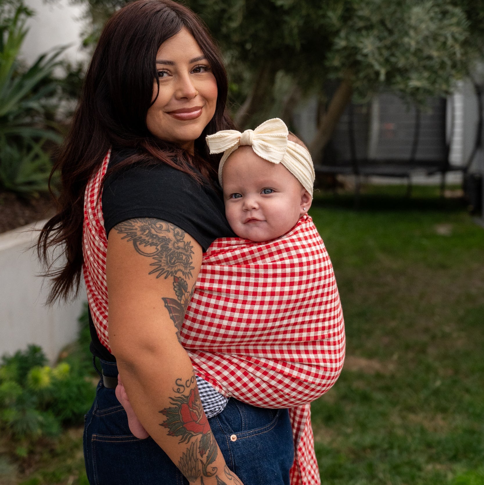 Woman holding a baby in a red and white gingham ring sling outdoors.