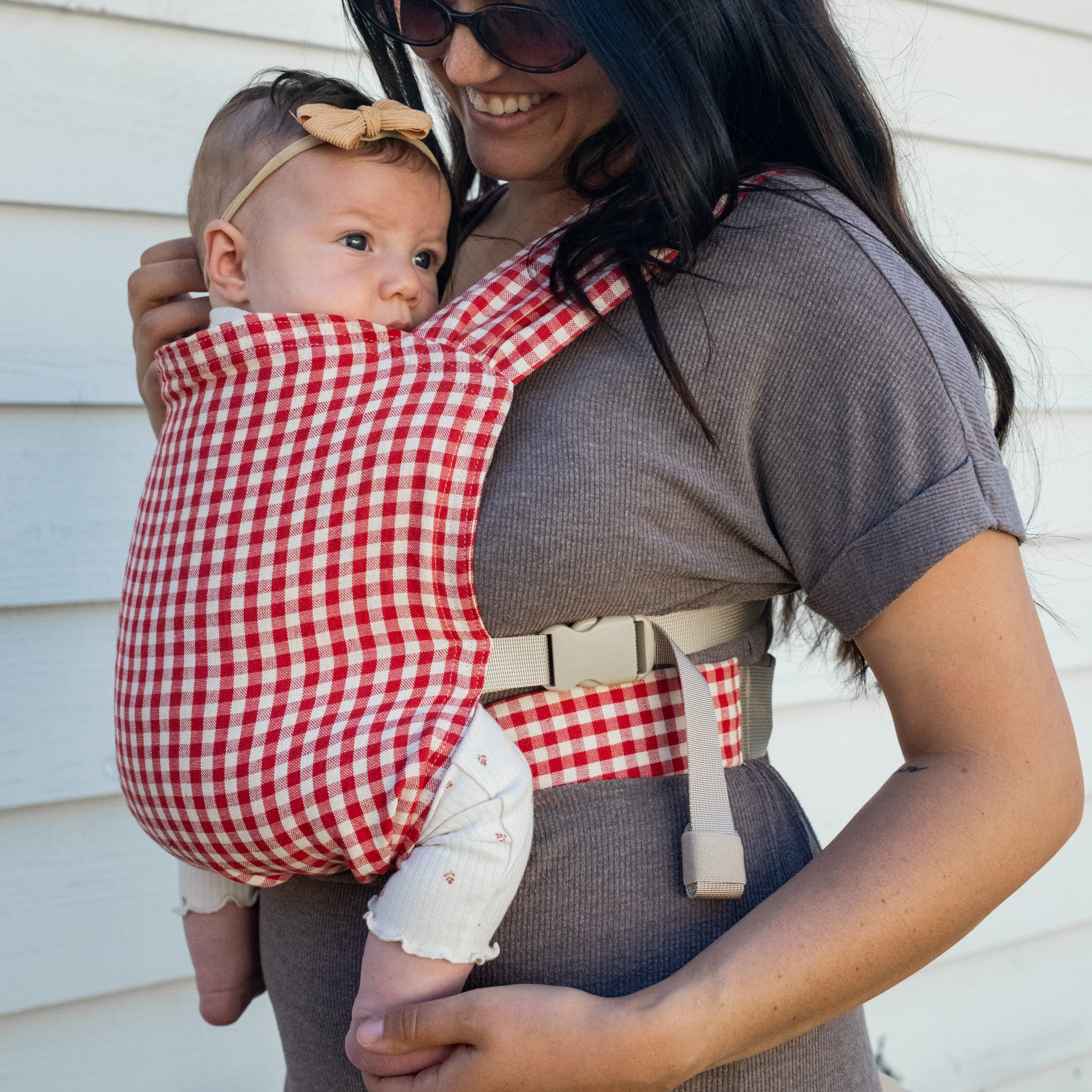 Woman carrying a baby in a red and white gingham buckle newborn carrier outdoors.
