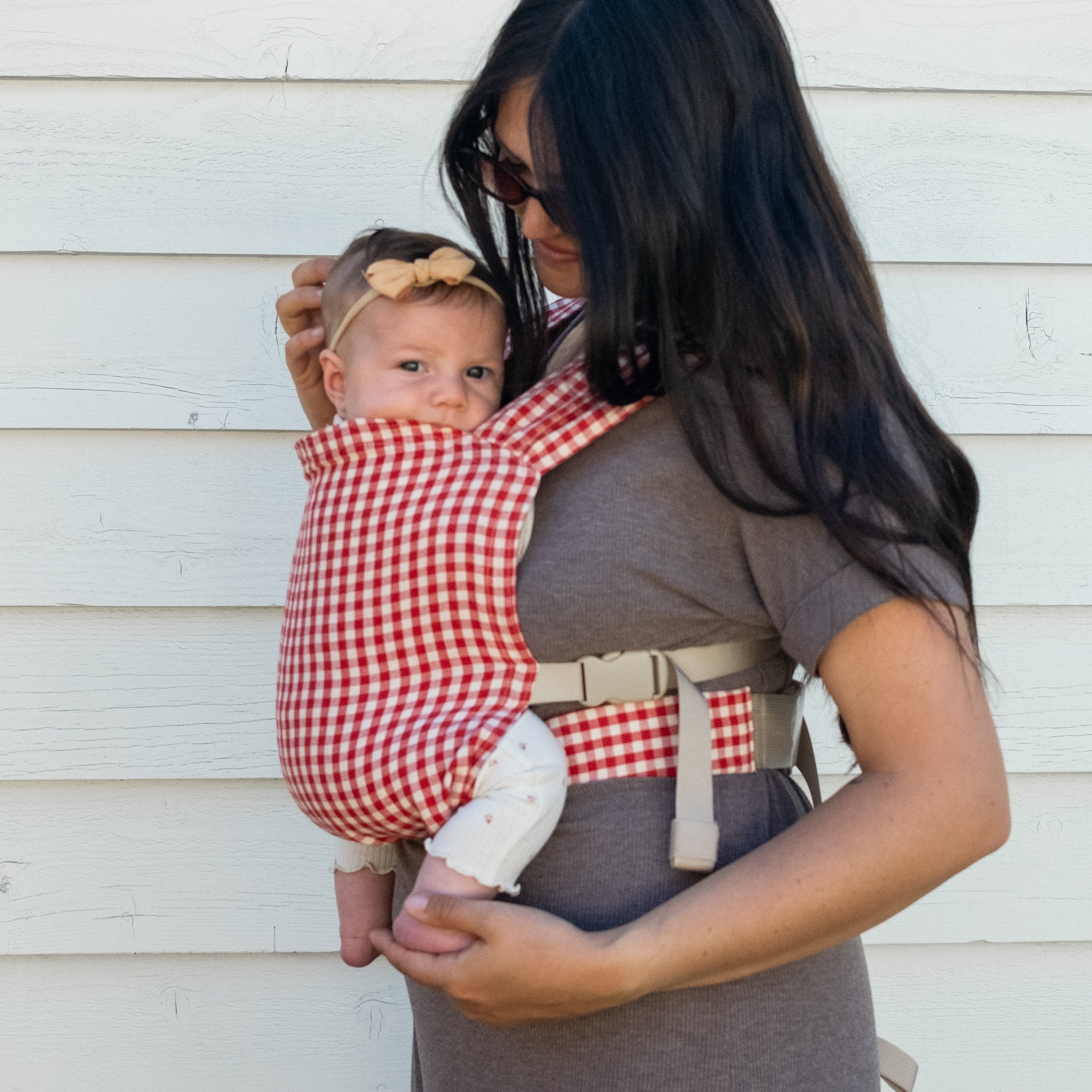 Woman holding a baby in a red and white gingham newborn buckle carrier against a light-colored wall.