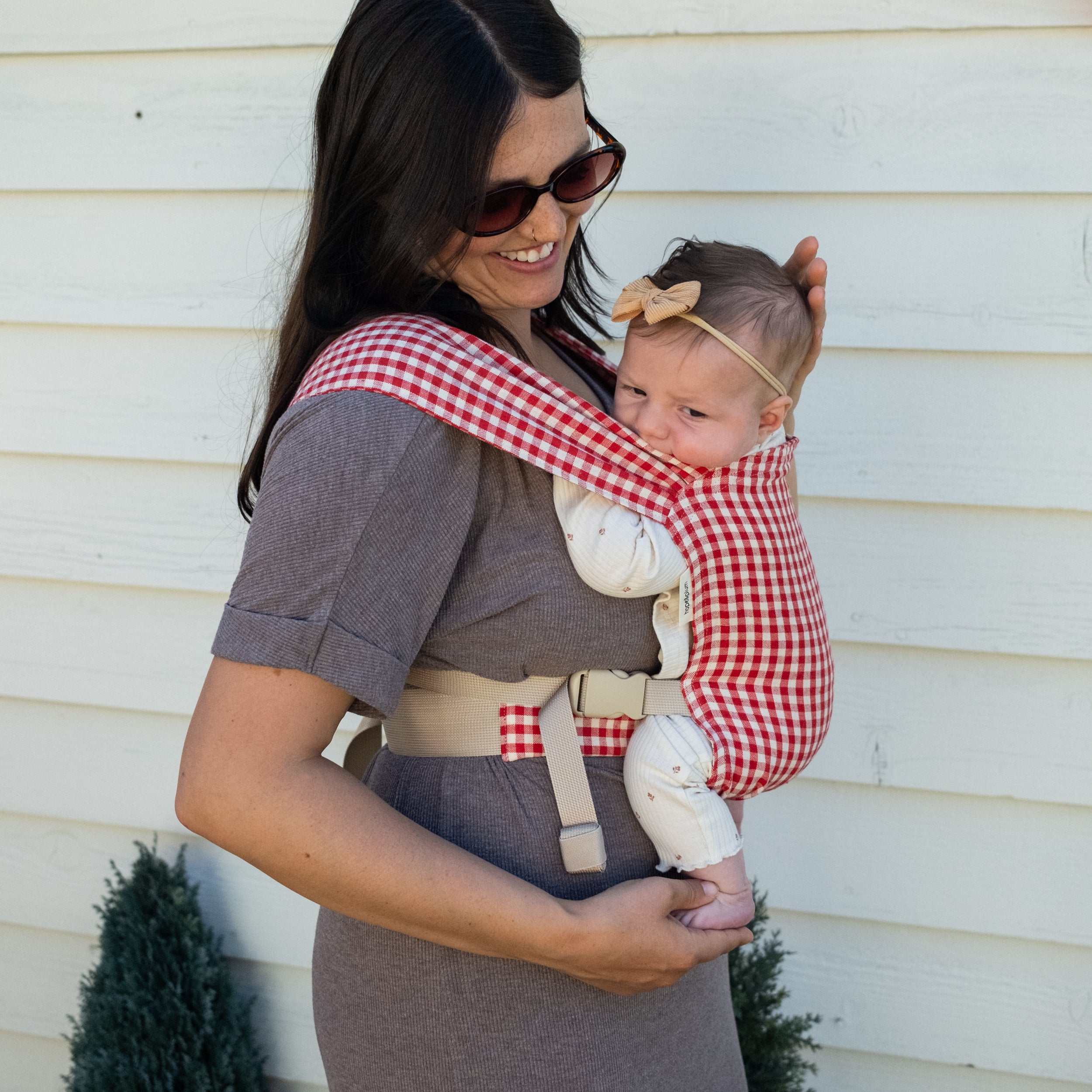 Woman holding a baby in a red and white gingham newborn buckle carrier against a light-colored wall.