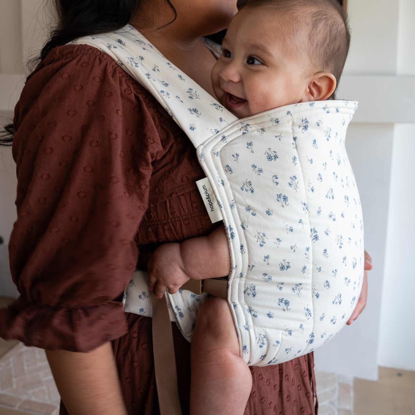 Woman holding a baby in a floral baby carrier indoors.