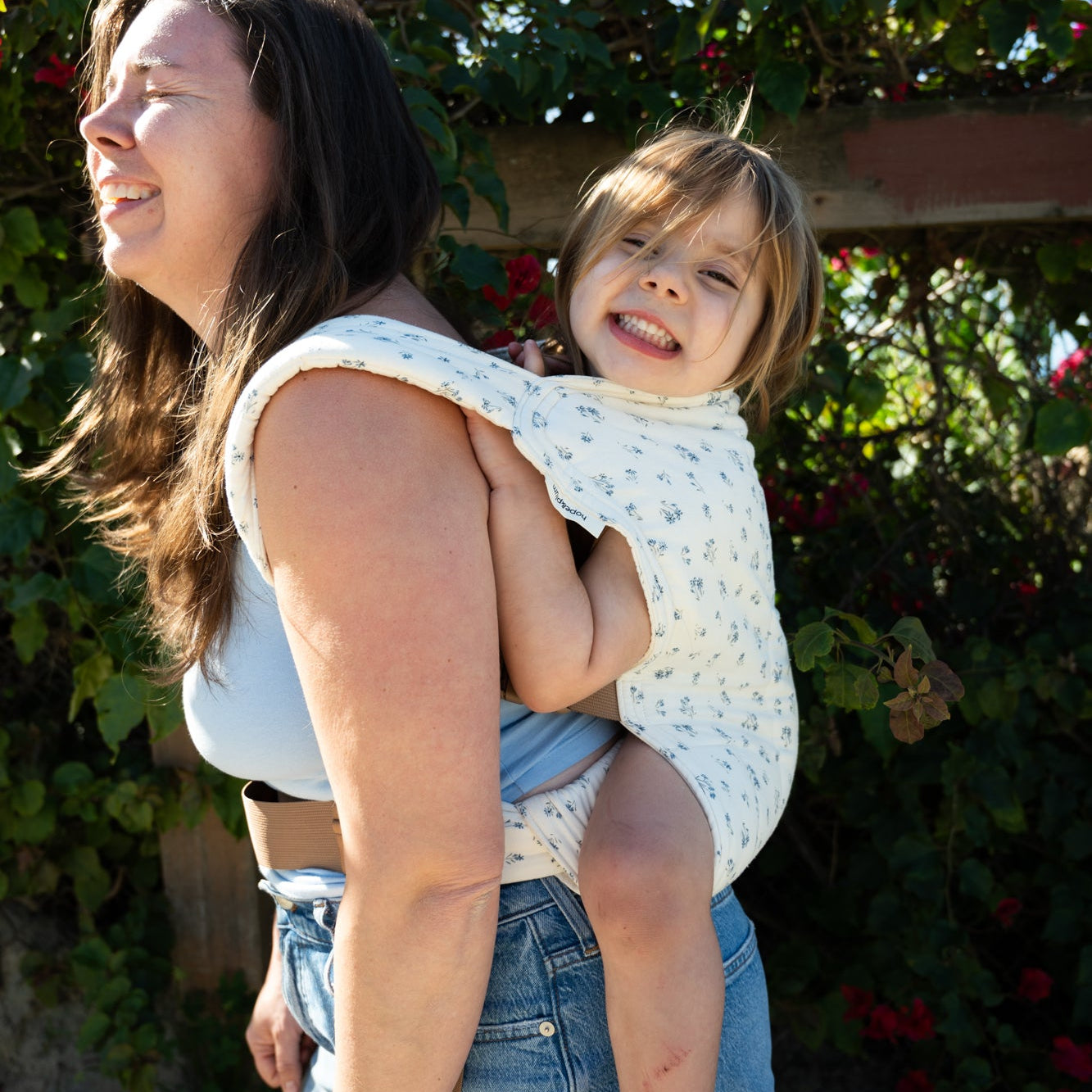 Woman holding a child outdoors in a light floral baby carrier with greenery and flowers in the background