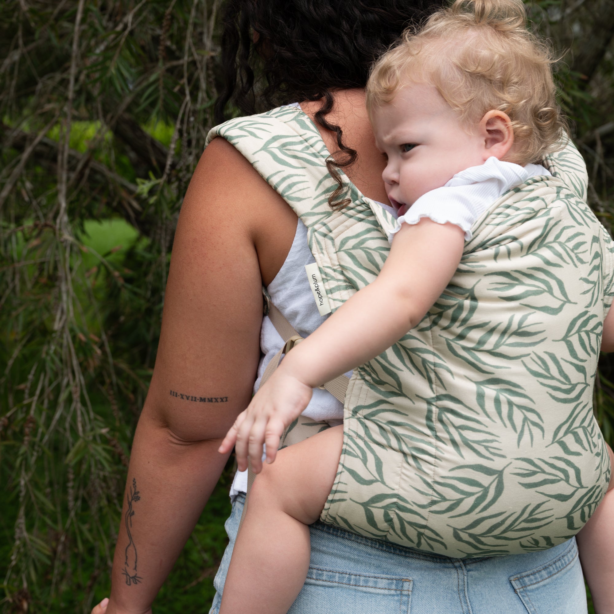 Woman holding a baby in a green leaf-patterned carrier outdoors.