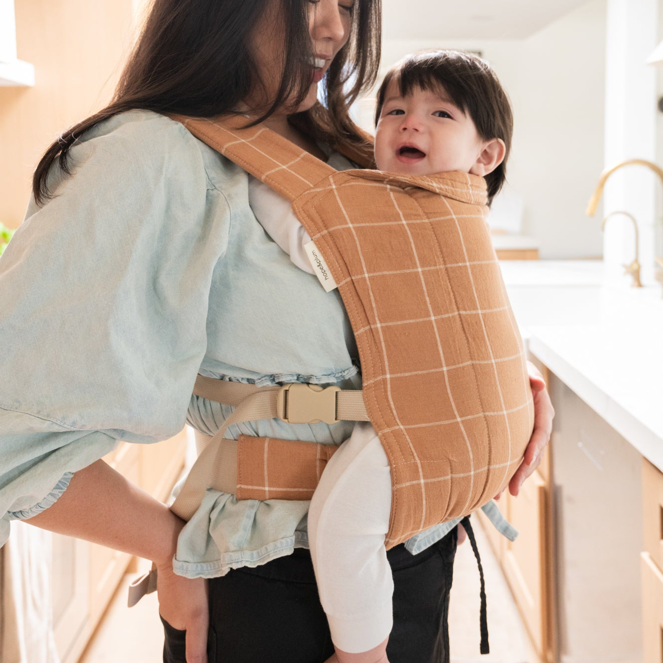 Woman holding a baby in a brown baby carrier with a white grid.