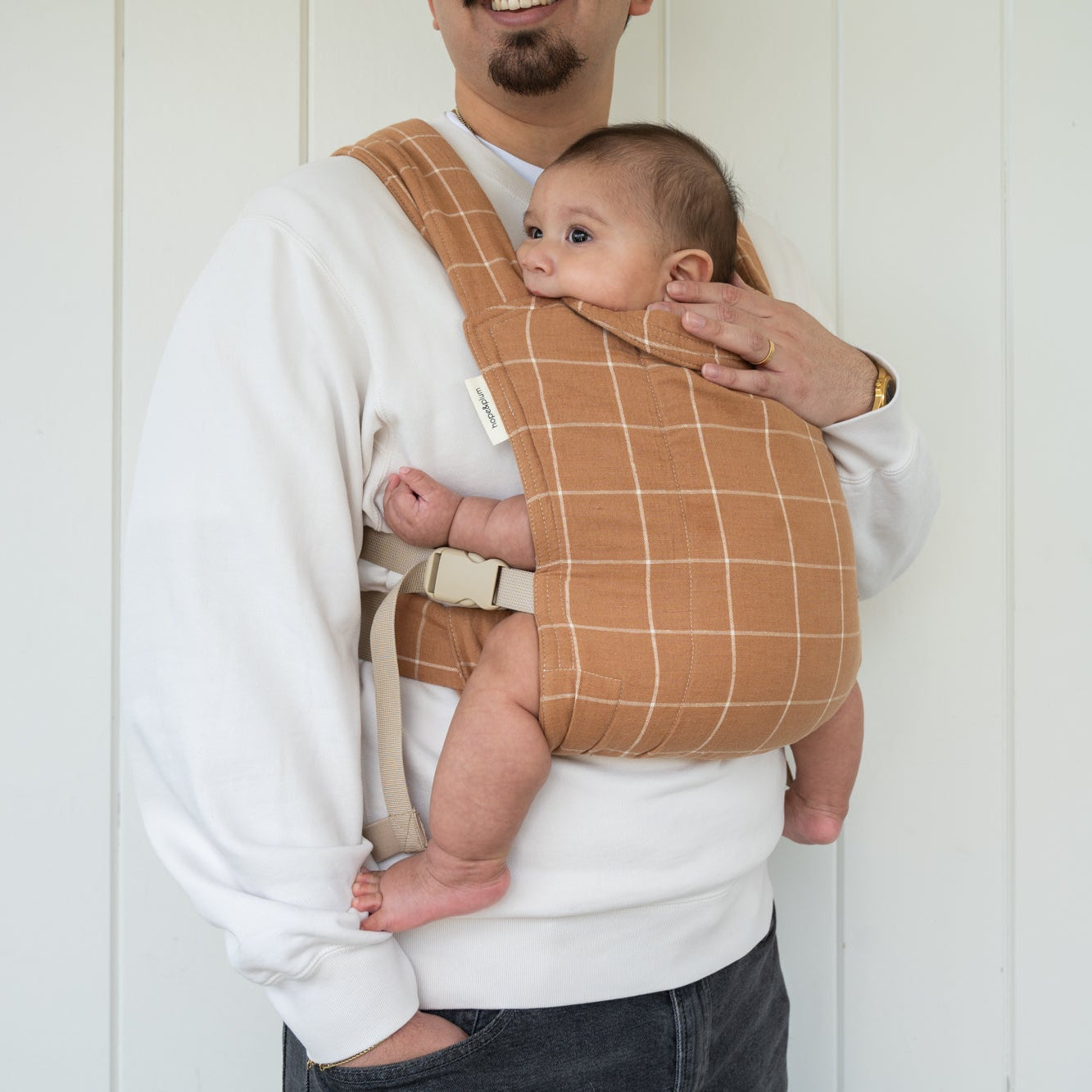 man holding a baby in a brown baby carrier with a white grid.