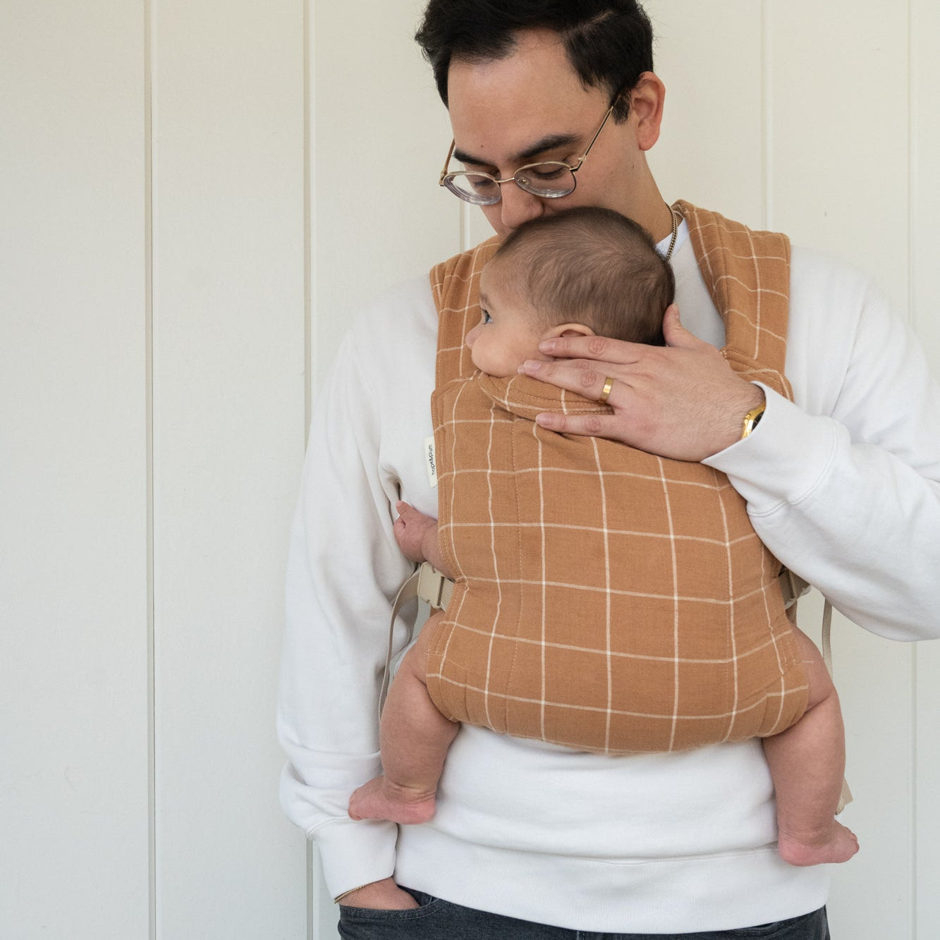 Man holding a baby in a brown grid-patterned carrier against a white wooden background